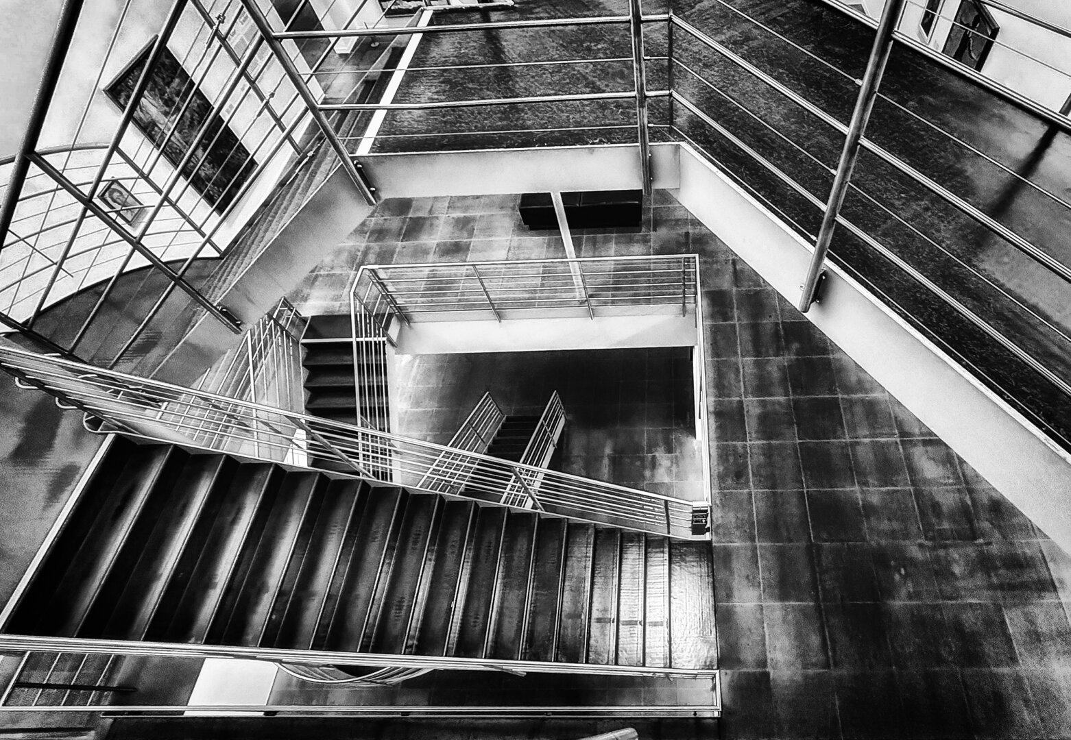 Eine kontrastreiche Schwarz-Weiss-Aufnahme blickt steil von oben hinab in ein mehrstöckiges, modernes Treppenhaus im Kunstmuseum Bern. Die geometrische Komposition wird durch klare Linien, rechteckige Treppenläufe und Metallgeländer bestimmt. Stufen und Absätze verschachteln sich in der Tiefe zu einem dynamischen, fast abstrakten Muster aus Licht, Schatten und dunklen Plattenbelägen. An einigen Wänden sind Kunstwerke zu erkennen.
