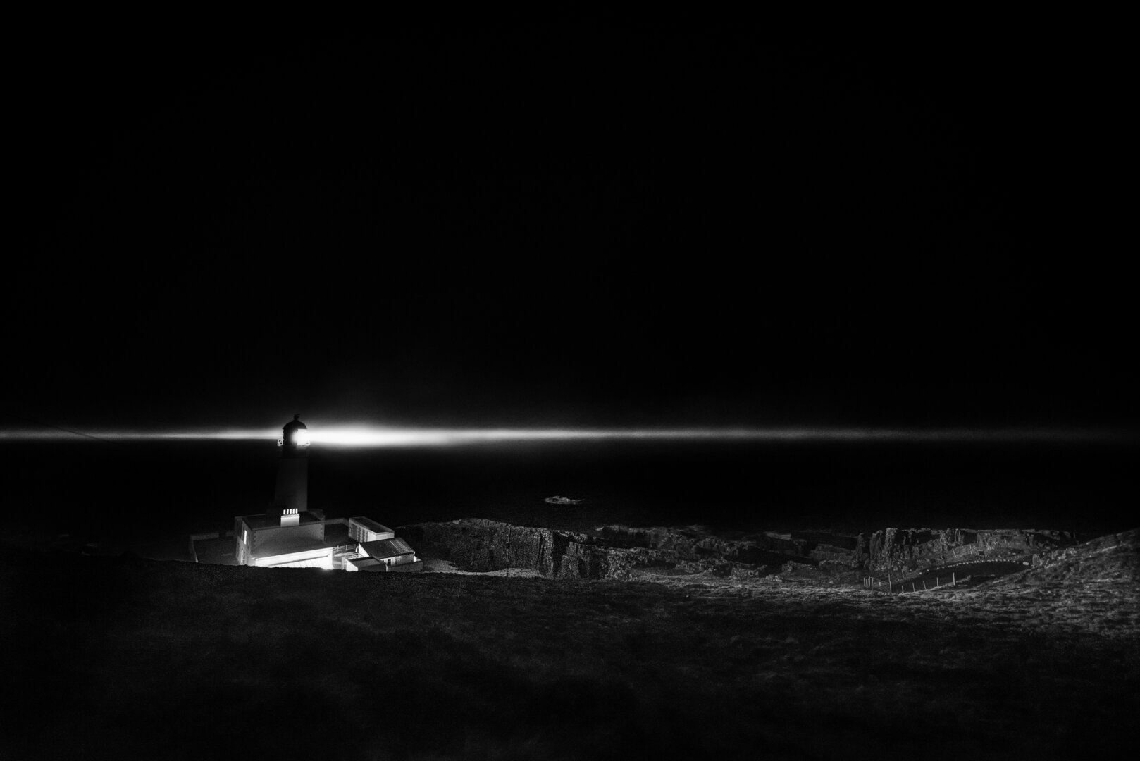 Nachtaufnahme eines Leuchtturms auf einer felsigen Steilküste, dessen Lichtstrahl waagrecht über das dunkle Meer streift; das beleuchtete Wohngebäude am Fuss des Turms hebt sich hell von der umgebenden Dunkelheit ab.