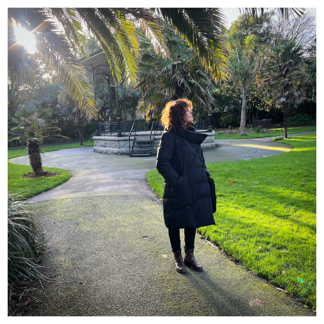 A coated female figure stands looking out of frame, lit from behind creating a halo of lit hair, under a palm tree.