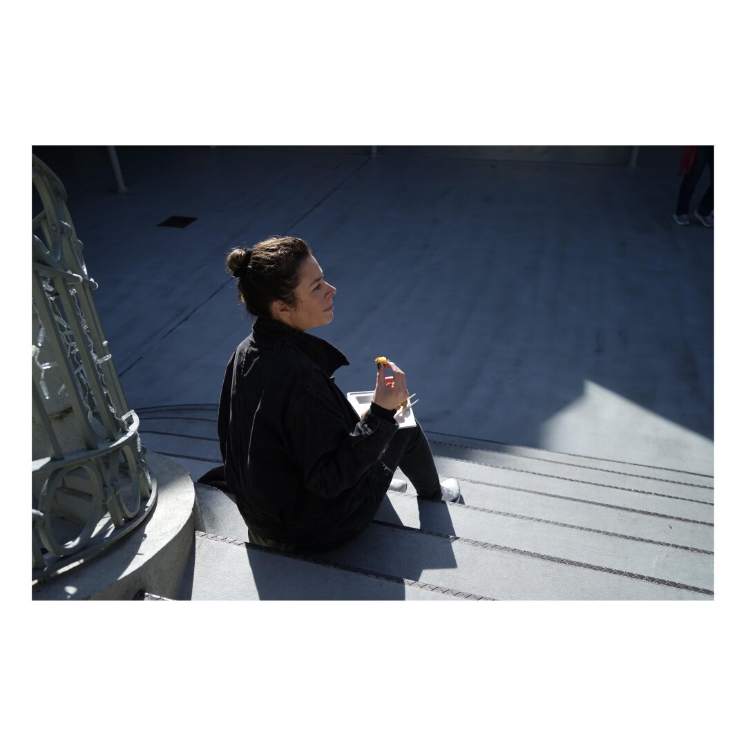 Viewed from above and behind a woman sits on steps eating prawn balls lit from high left with a contrasting shadow behind her.