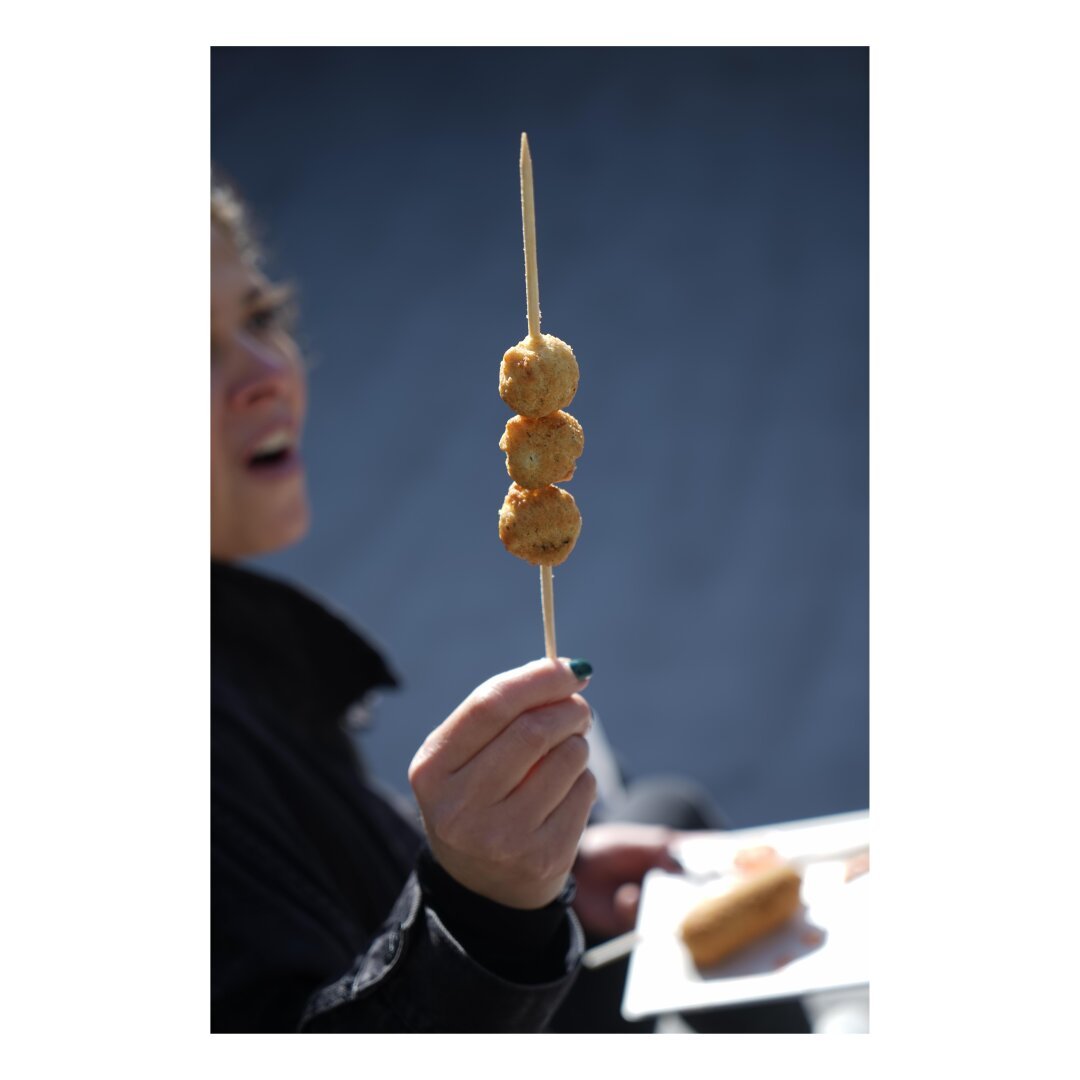 Woman holds three prawn balls on a skewer up as she chews through a hot one out of focus frame left.