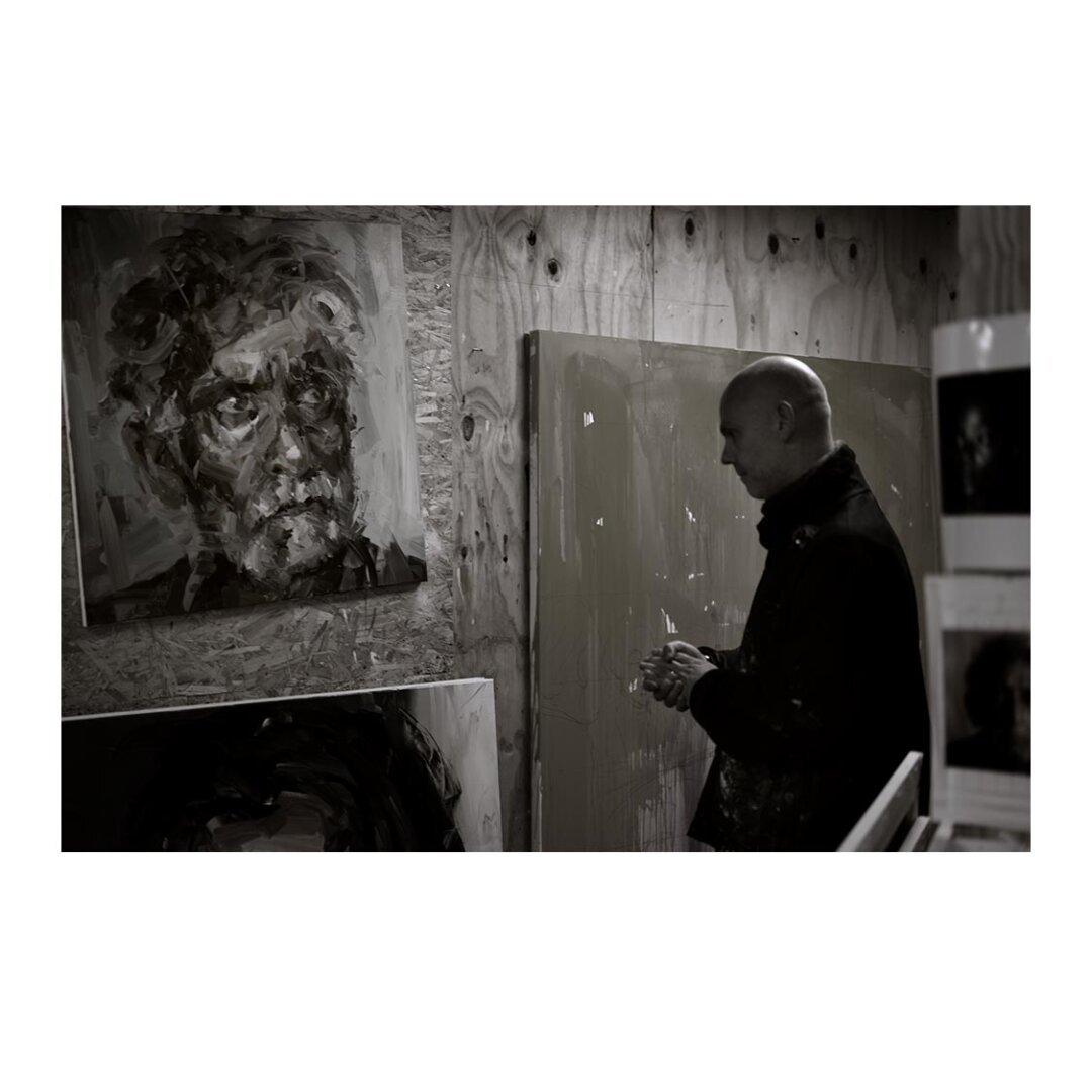 [imnage is black and white] a man (Artist Paul Wright) clad in paint covered clothes, stands frame-right hands clasped looking thoughfully to frame-left. A large painted older man's head [frame-left] looks back at Paul.