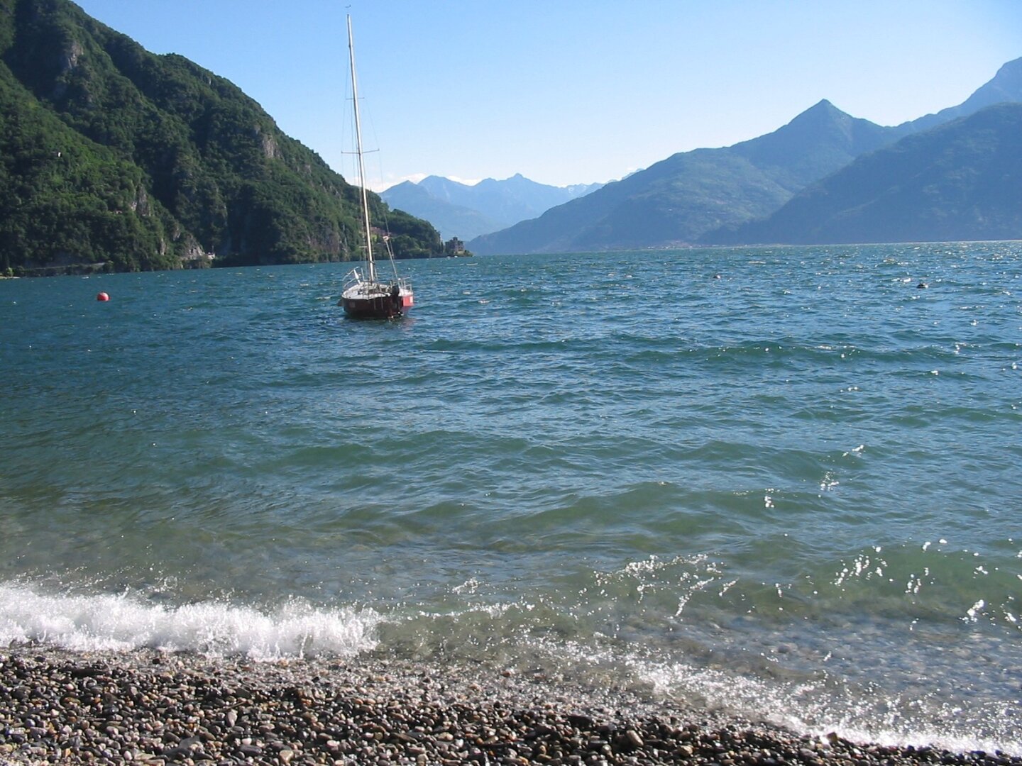 Lake Como, mountains in the background, waves in the front crash on a small pebble beach. A sailboat moored off  in the centre left