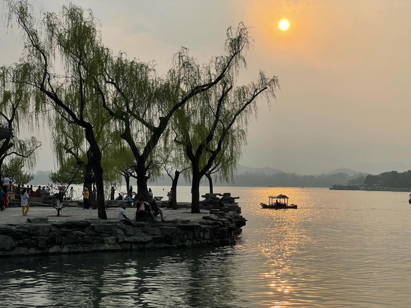 A picture of a boat on a lake with the sun behind.  There are trees on land near the boat in the foreground.