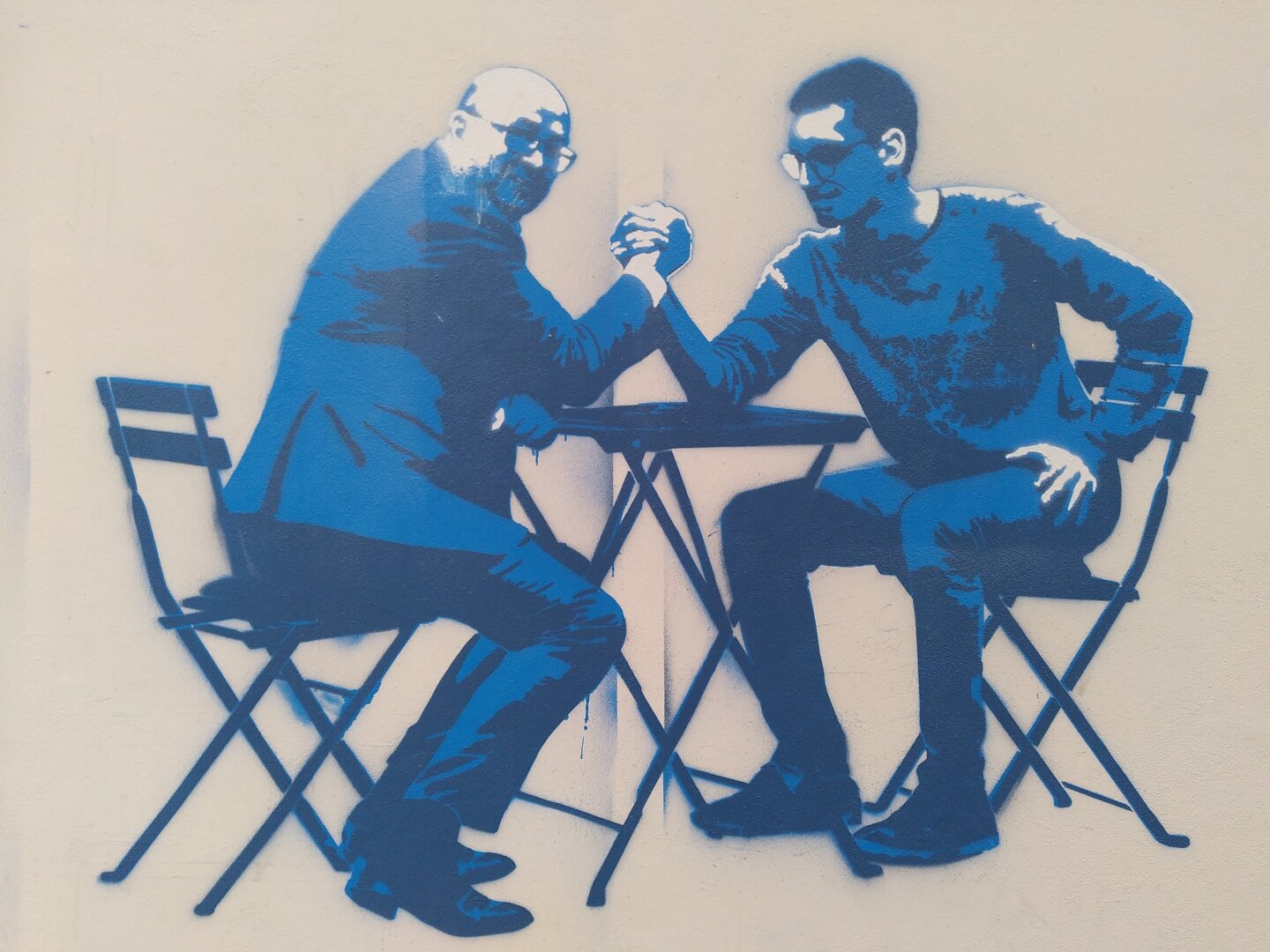 Two men, a young and an older one sit on chairs opposite of each other. In between is a table and they do arm-wrestling.