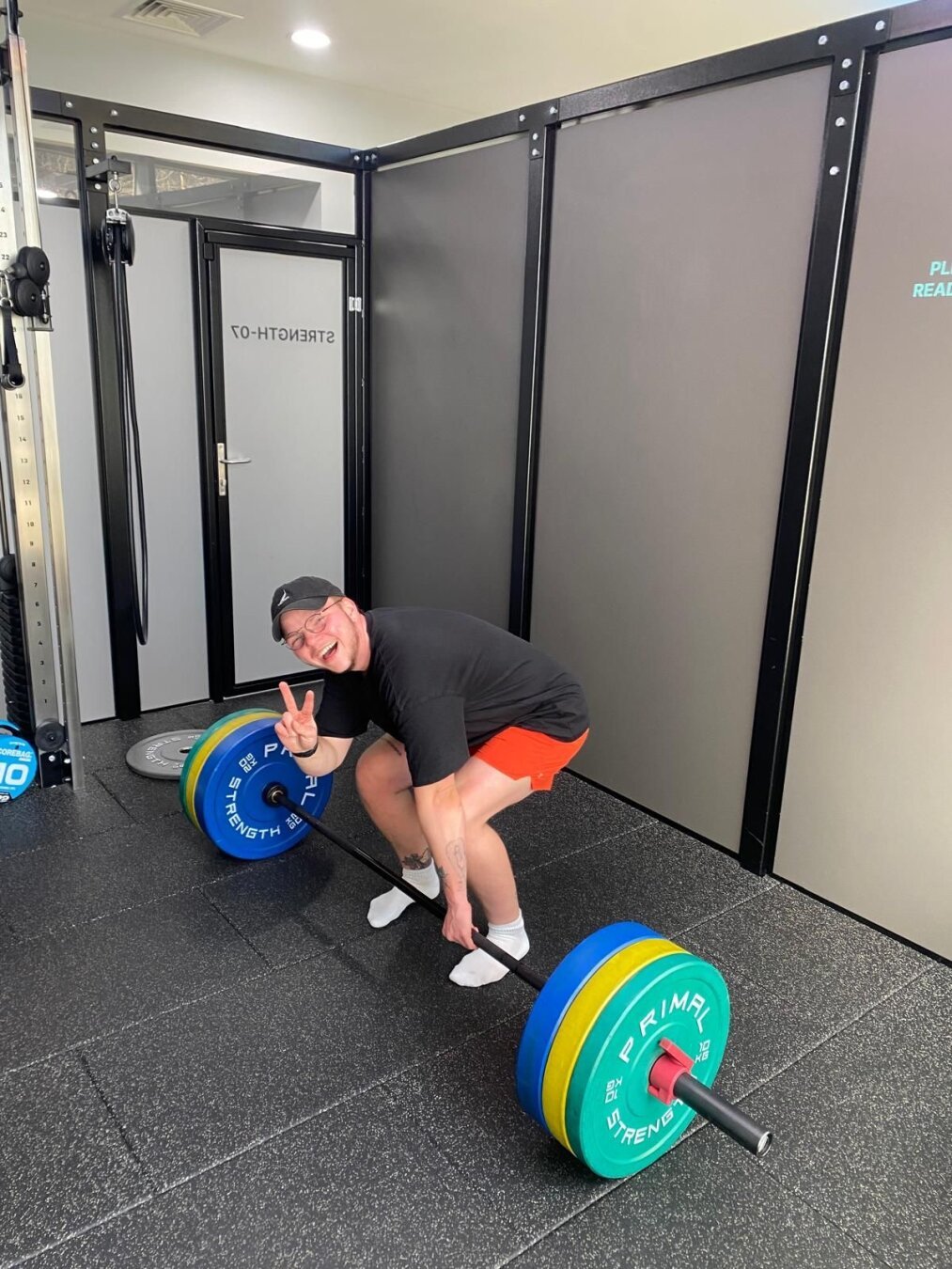 A white man with a black hat, orange shorts and black t-shirt in the gym, giving the peace sign while holding a barbell with 110kg in plates on it.