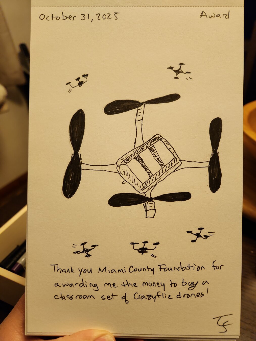 Pen sketch of a Crazyflie drone (with horrible perspective, whoops!) This is a little quadcopter that fits in your hand. There are little silhouettes of five other drones with whoosh lines to indicate movement. The caption reads "Thank you Miami County Foundation for awarding me the money to buy a classroom set of Crazyflie drones!"