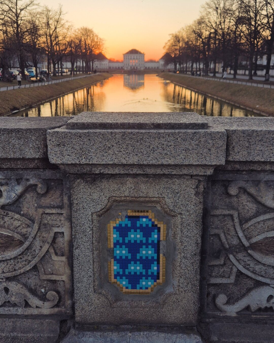 Sundown at Nymphenburg castle, redish sky and a tree-lined canal reflecting in the water with the castle at the end of the canal and a part of the stone bridge with a space-invaders inspired street art in the foreground