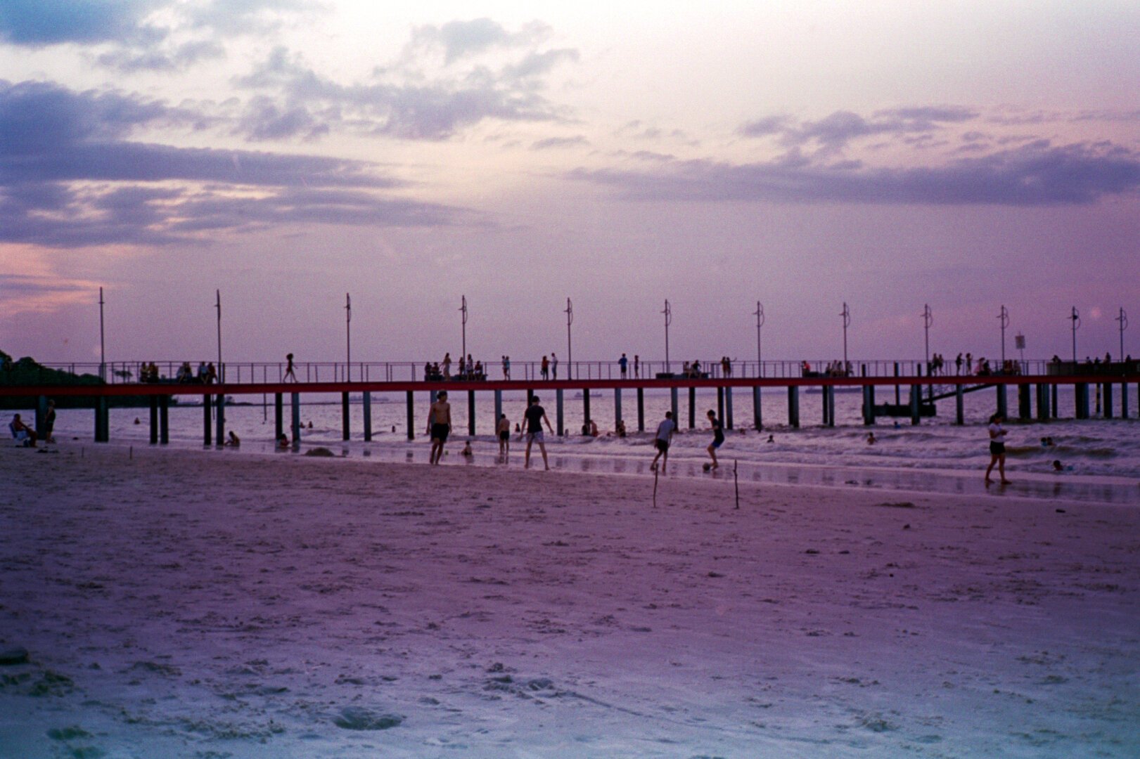 A scenic view of a pier extending into the ocean at dusk. Silhouettes of people are scattered along the pier and the beach. The sky is a blend of soft purples and blues, with fluffy clouds scattered above.