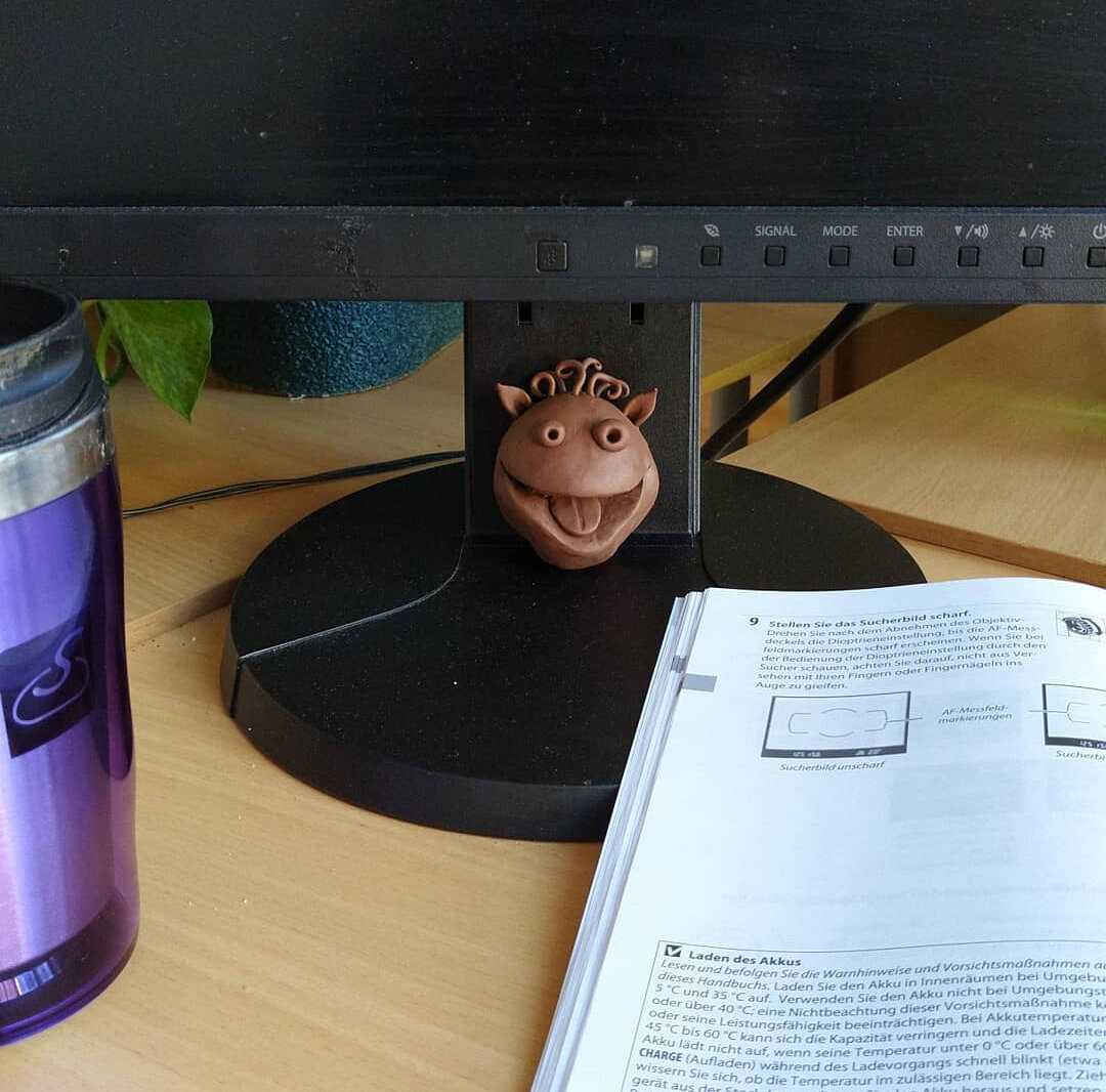 A workspace scene showing the same whimsical plasticine creature sculpture from the previous image, now positioned on a black circular platform or turntable. The sculpture sits in front of a computer monitor, surrounded by various items including a purple water bottle or tumbler on the left, an open German instruction manual or book on the right, and what appears to be a green leaf and blue textured object in the background. The setting appears to be a desk or workstation with wooden surfaces.