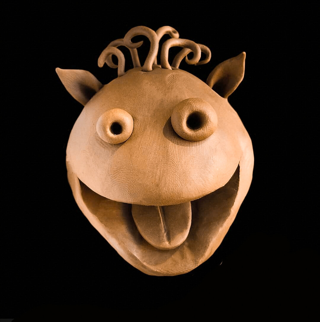 A whimsical plasticine sculpture of a cartoon-like creature's head against a black background. The figure has large, bulging round eyes, small pointed ears, curly hair or tentacles sprouting from the top of its head, and an oversized open mouth with a protruding tongue, giving it a playful and slightly goofy expression.