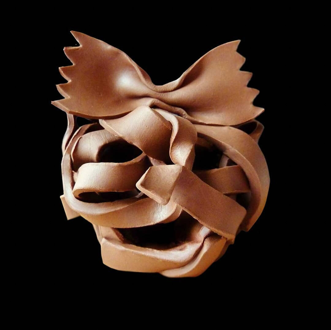 This image shows an artistic plasticine sculpture meticulously crafted to resemble pasta, specifically farfalle (bow tie or butterfly pasta). The brown plasticine has been skillfully manipulated into thin, ribbon-like strips with jagged edges along the outer portions, mimicking the characteristic shape of the pasta. The clay model features multiple layers with elegant folds and curves, creating a three-dimensional representation against a black background. The plasticine has a smooth, slightly glossy finish that highlights the detailed craftsmanship.