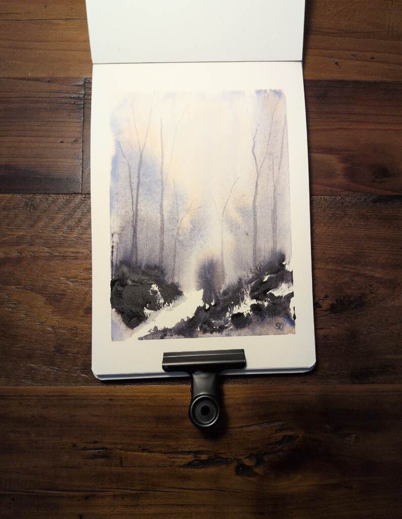 This is an Atmospheric Watercolor Landscape Painting in portrait orientation in an A5 sketchbook using Schmincke Tundra Violet.  The dark masstone makes up the terrain with a little dilution and granulation and peaks of white paper. Above the horizon line, Tundra Violet is diluted creating bursts of warm granulating blue and a halo of warm pale brown. Thin leafless trees stretch up from the horizon line.The overall feeling is that of a frosty fall morning.