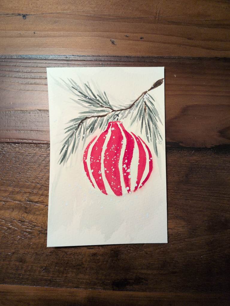 This is watercolor holiday card in portrait orientation on a 4x6 sheet of Arches Coldpress watercolor paper.  For this card I painted a branch of an evergreen tree with a peppermint inspired bauble hanging from it.  The painting is splattered with white paint to kind of give the appearance of snow.