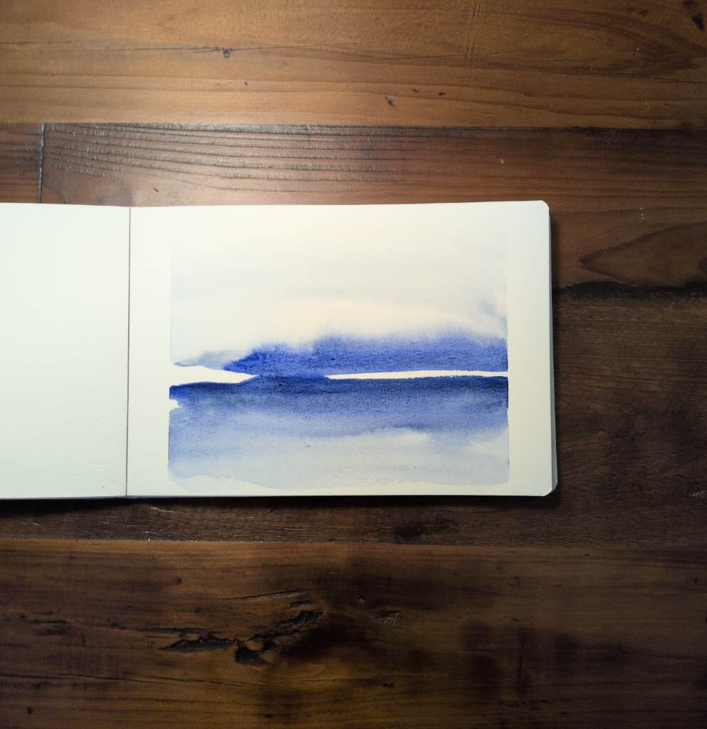 This is a horizontal atmospheric watercolor landscape painting in landscape orientation in an A5 sketchbook. Using only horizontal brushstrokes this Atmospheric Landscape Painting is done with French Ultramarine and a hint of Neutral Tint at the horizon line. There is a strip of white at the horizon that is interrupted by a color bleed towards the left side. The colors bleed and granulate and are stronger below the horizon line and much more diluted above the horizon line.