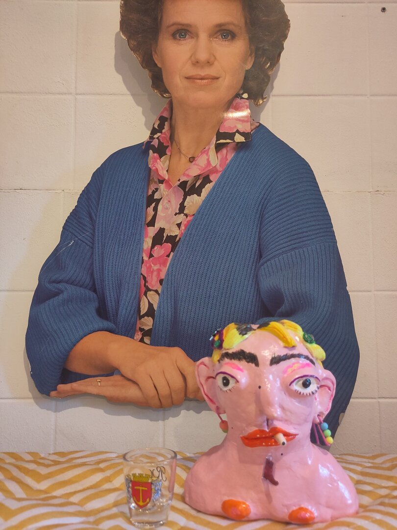 A head made from air drying clay. She has blonde hair and a stoic expression. The torso of another woman wearing a blue west is behind her while she's smoking nakedly.