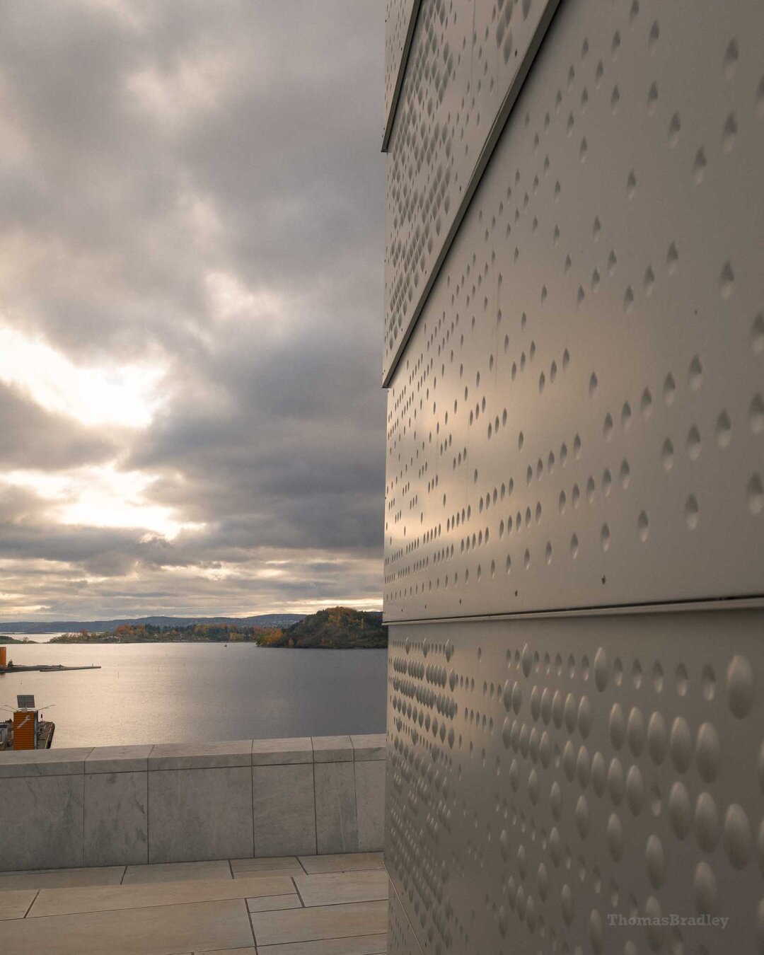 A view from the rooftop of the Oslo Opera House. The image is split by a modern metal wall dotted with round, Braille-like indentations. To the left, soft golden light breaks through dramatic gray clouds over a still fjord, with forested land in the distance and marble tiles in the foreground.