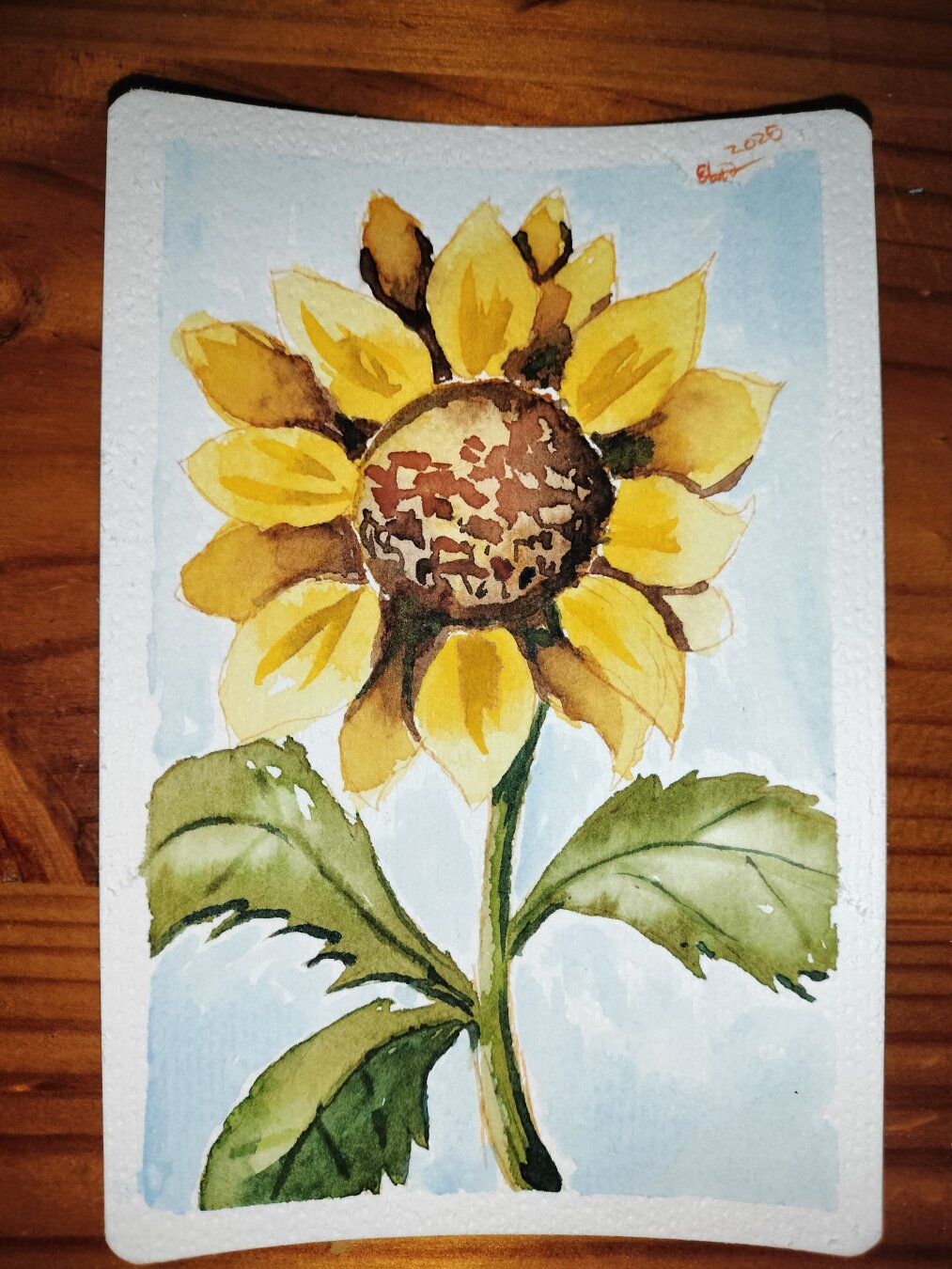 A watercolor painting of a bright yellow sunflower, it has three green leaves and a pastel blue background.