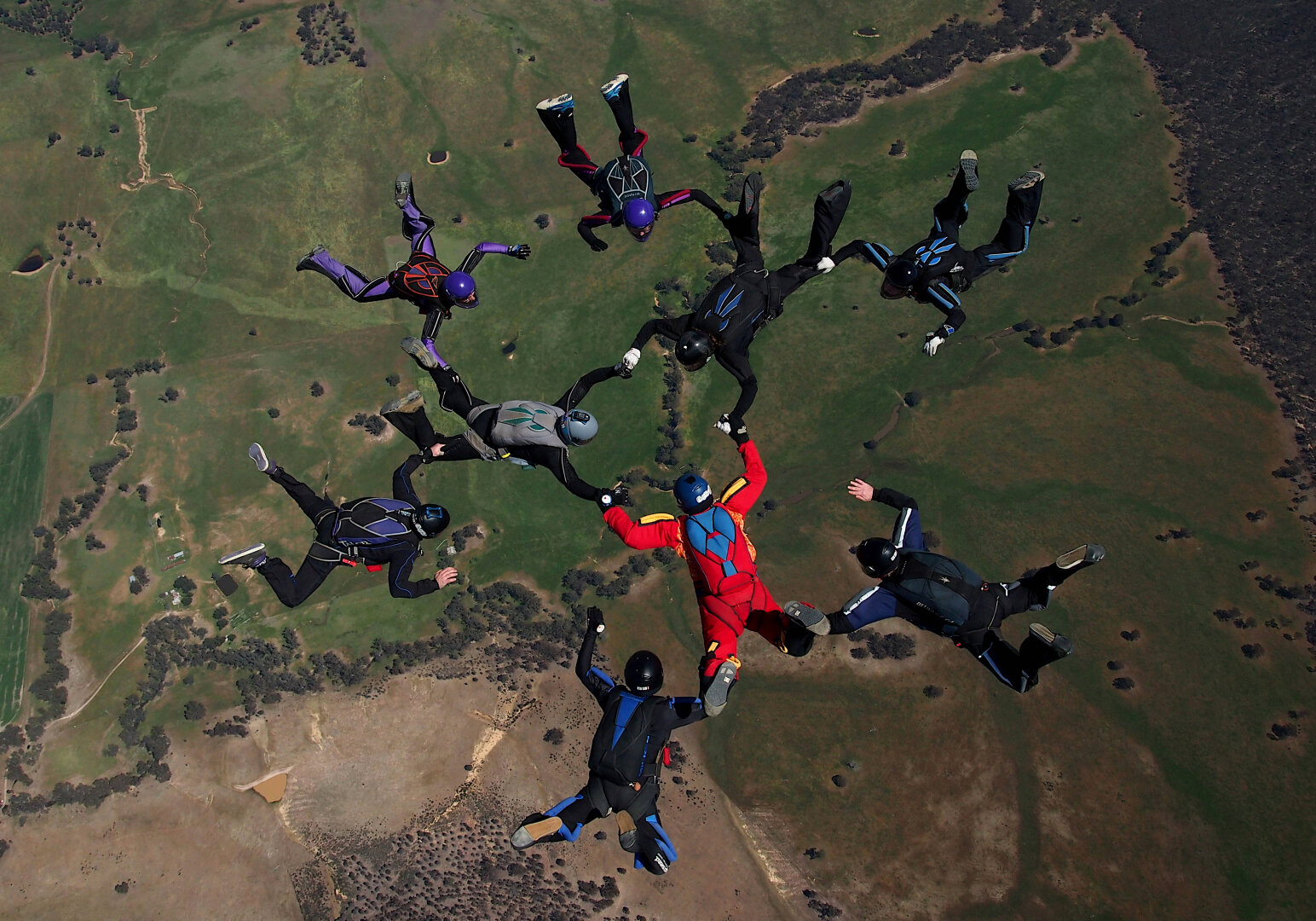 Image of nine people in a freefall formation taken from above the formation. The earth below is a browny green with dry patches and marked with areas of tempory water flow. Image taken in Australia
