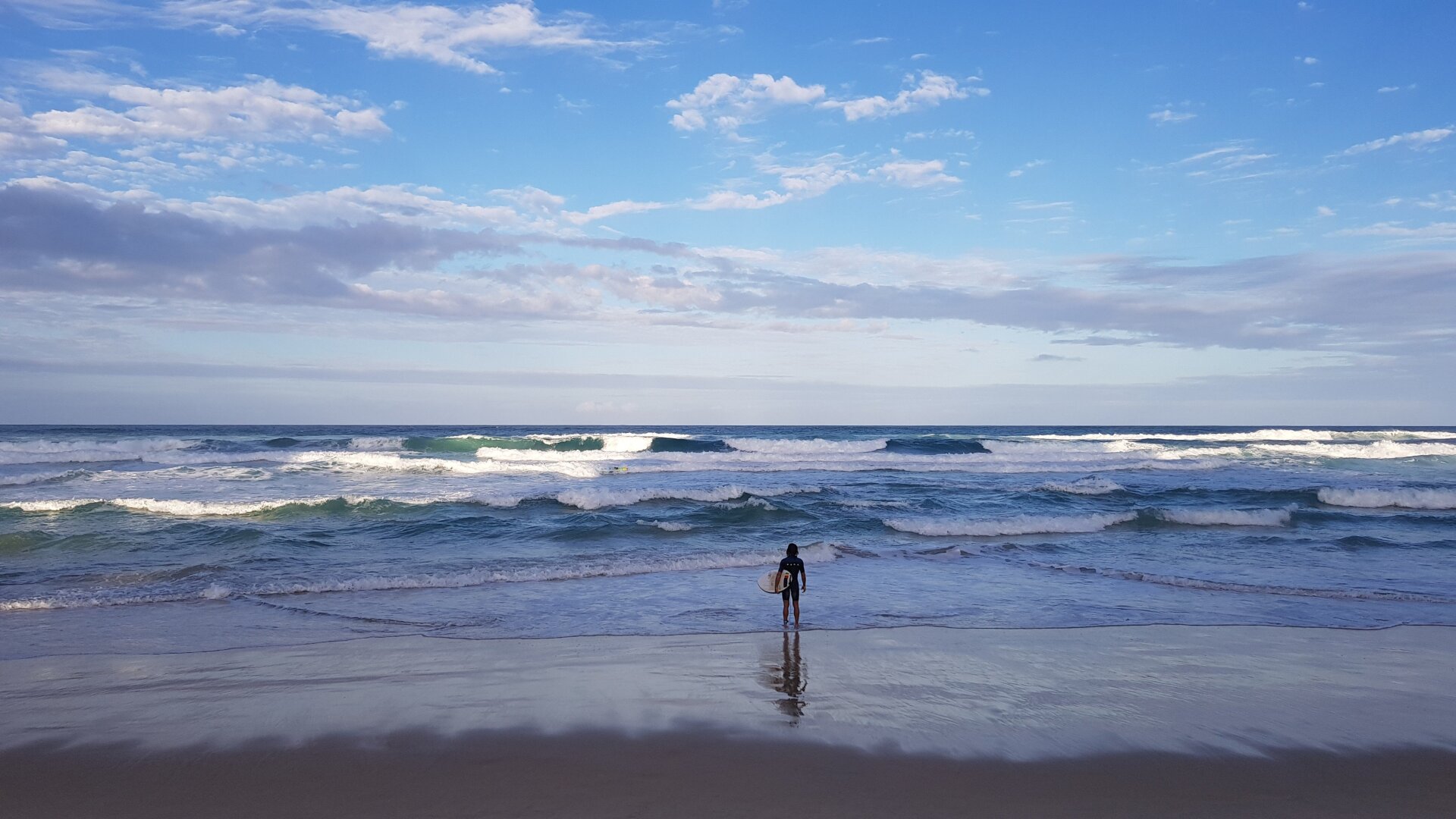 A lone surfer standing on the shoreline looks out to sea at the breaking waves.