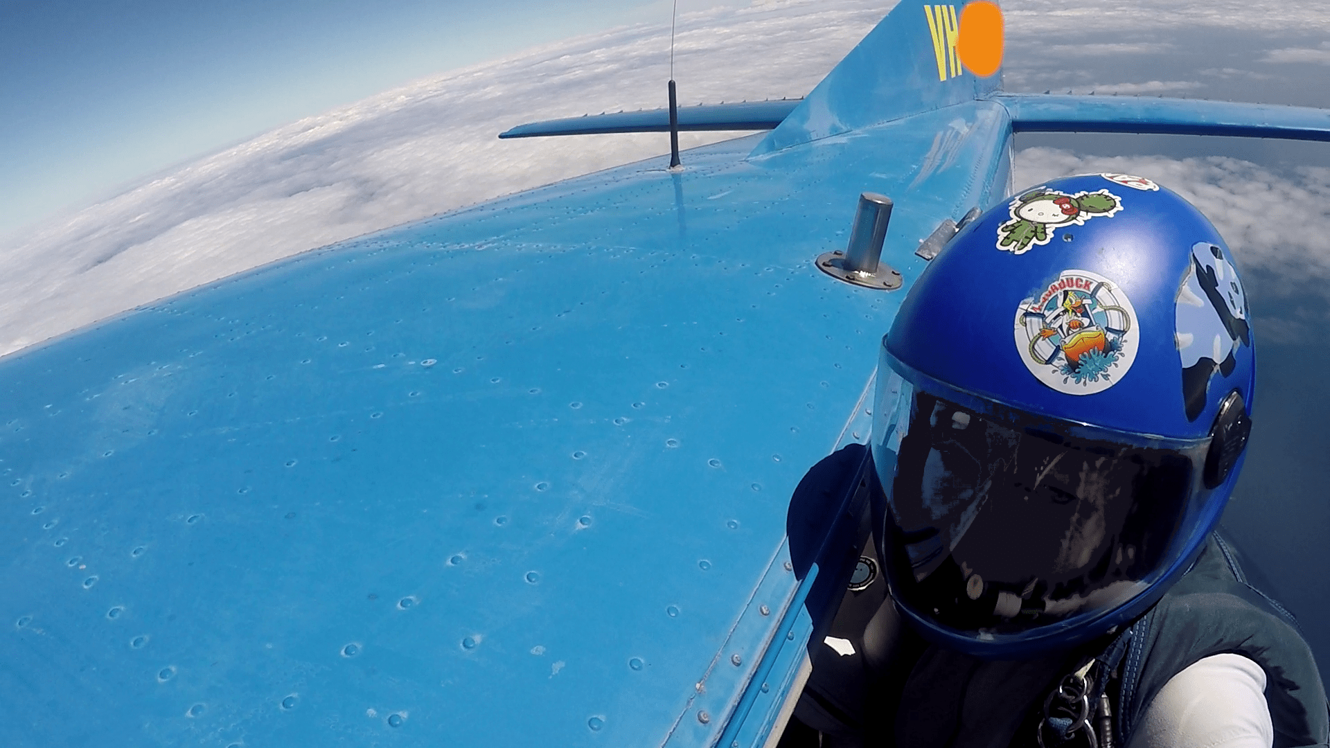 A girl skydiver hangs from the rear step of a blue jump aircraft. She's looking at the camera man. She is wearing a blue full face helmet with cute animal stickers, a duck, a panda and some unknown character.

Over the top of the aircraft a large bank of cloud can be seen in the distance behind the the aircraft's vertical stabilizer.