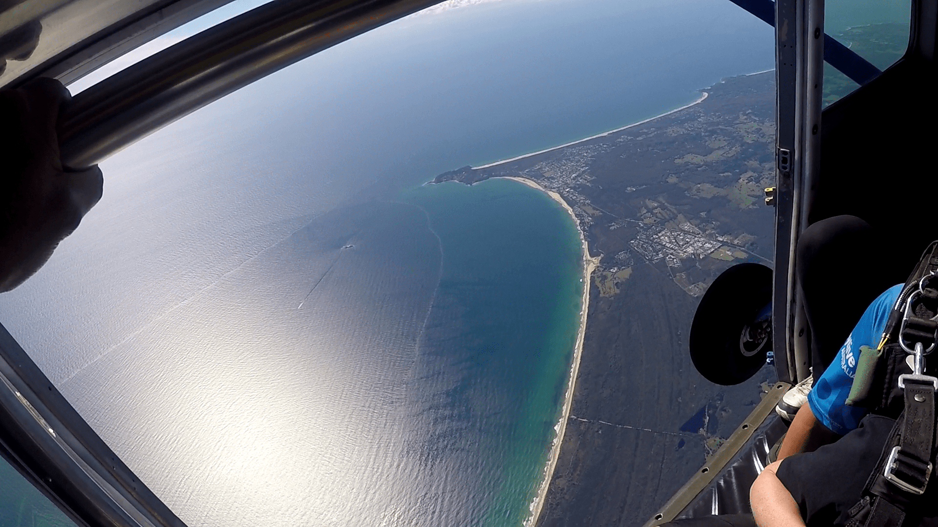 Image taken from inside a jump aircraft on jump run. The door is open. Just inside sitting along edge of the door part of a tandem instructor and passenger pair is visible. Their faces cannot be seen. Above and inside the open door is a metal bar and the photographers hand. Out side the aircrafts left side wheel can be seen in the frame. Far outside, the view is of a glorious clear summer day. below is the South Pacific Ocean's blue/green water and the coastline leading to Cape Byron and the township of Byron bay. To the North and South of the town is forested up to the sandy beach that marks the coastline.