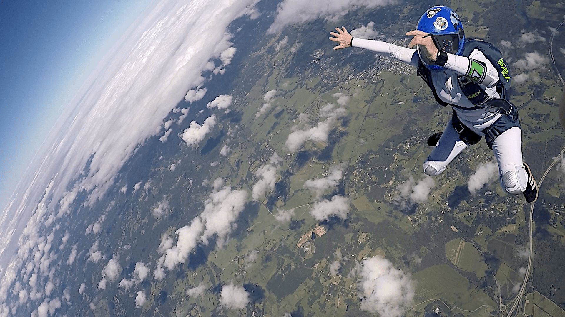 In the top right is a girl skydiver in blue helmet, white jumpsuit and her body is in a knee fly position. In the back ground far below and to the left are are small white cotton dotted over the green landscape. In far distance is a large front of sold cloud moving toward the dropzone.