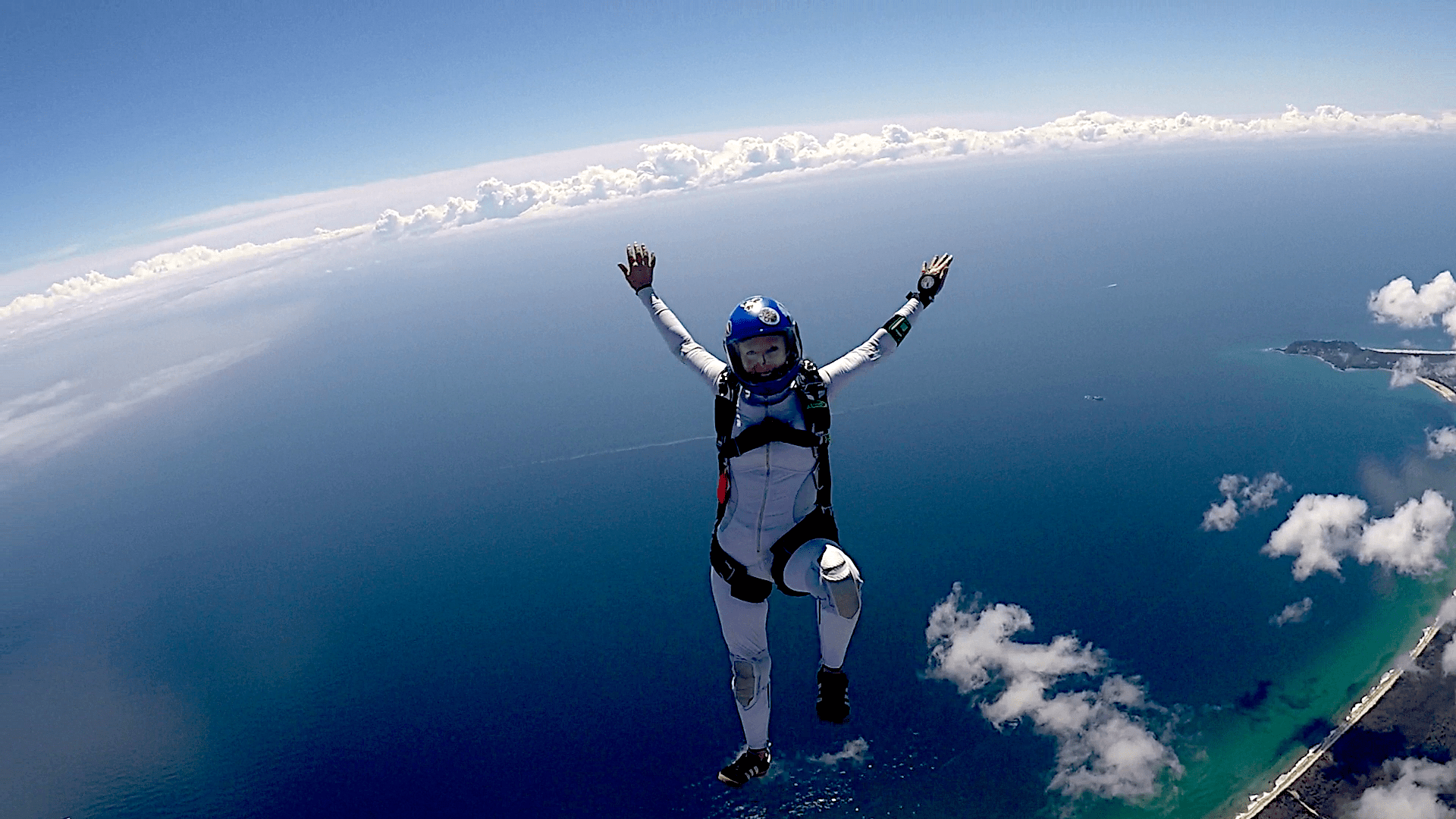 In freefall looking out at the wast blue South Pacific Ocean. In the foreground center a girl skydiver falls in a head up position. She is wearing a blue helmet and white jumpsuit. She is smiling.

In the far back ground over the ocean is a long narrow band of cloud the spans the length of the photo.
