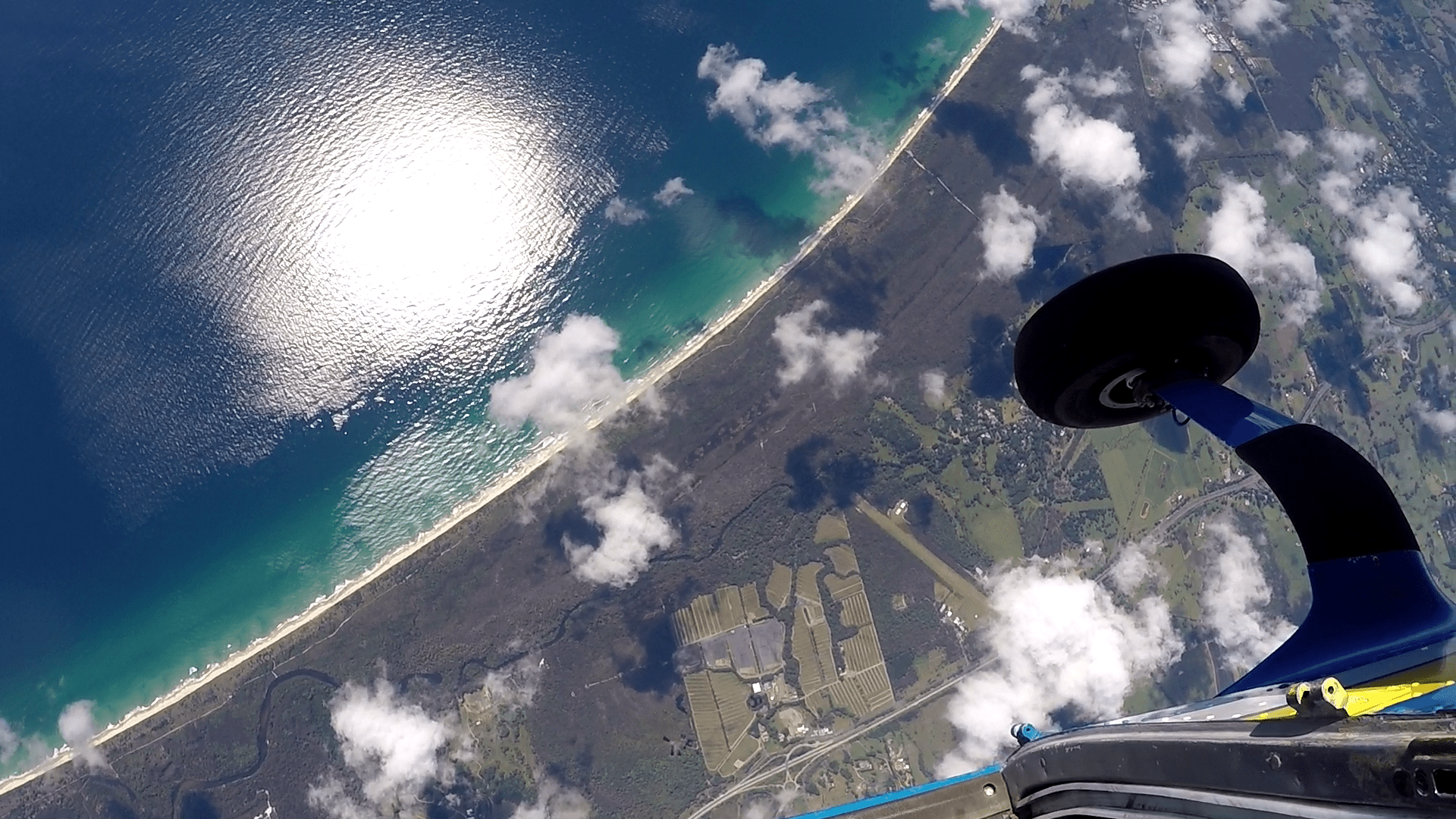Image is taken from inside the door of a jump aircraft on jump run and seconds before climb out looking directly down over the airport's grass runway. The airport is surrounded by a blanket of large gum trees. to the west past of the coast line is in the picture. Well below exit height the picture is dotted with fluffy white cotton ball clouds that mark the leading edge of an approaching cloud front.