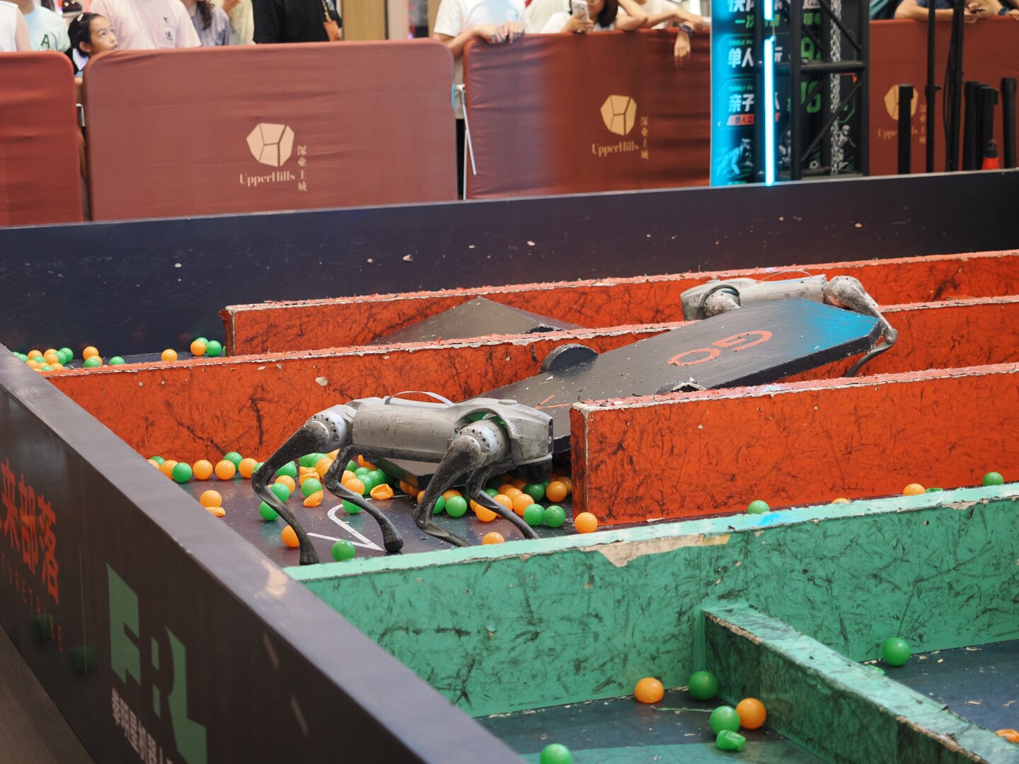 Two grey dog-style robots in some kind of maze or obstacle course. The outer wall is black and the inside walls that need to be navigated are green and red. The maze has ball pit style balls on the ground, a pivoting board, and some angled ground to walk over