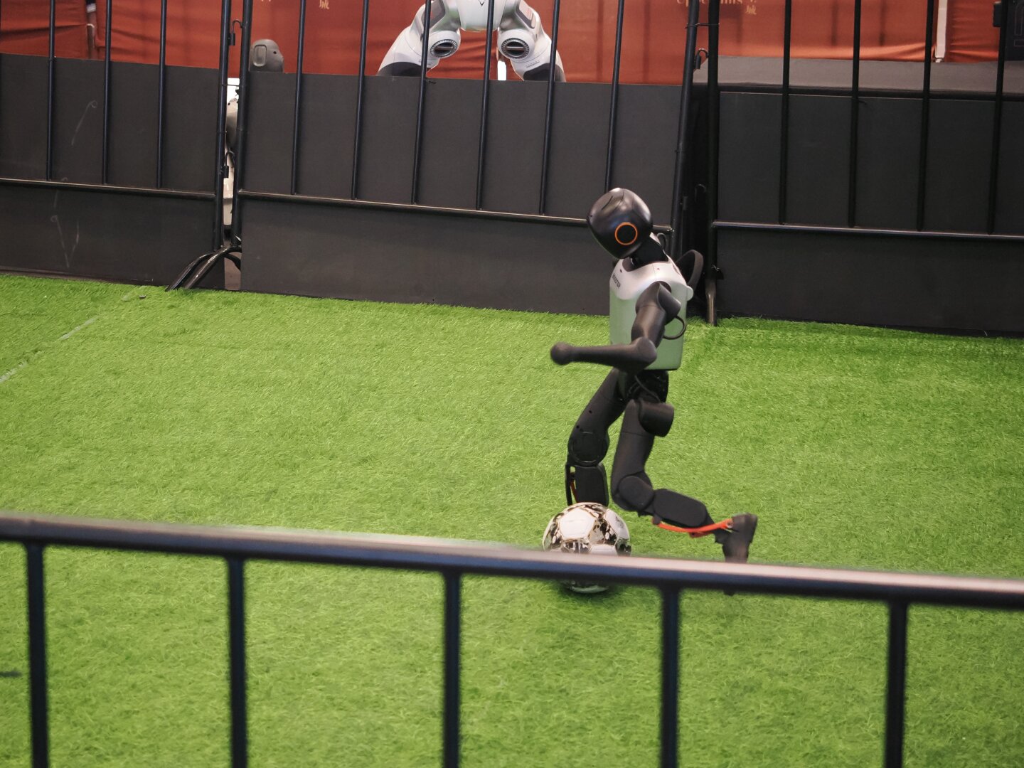 A small robot kicks a soccer ball behind a low railing