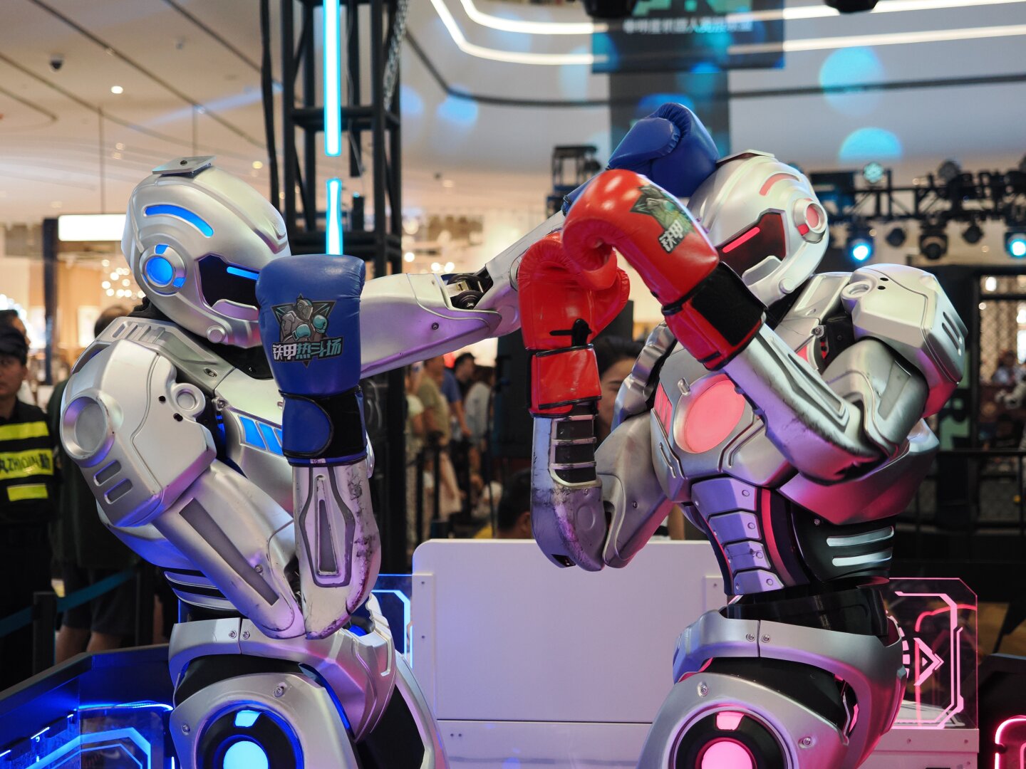 Two robots, one with red accents and red boxing gloves and one with blue accents and blue boxing gloves face each other. The blue accented robot throws a big left punch