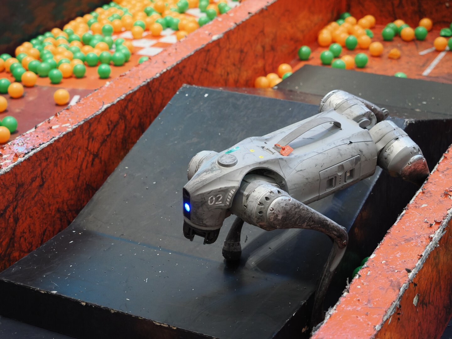 A dog-style robot stands on a sloping piece of black ground between two red walls. Around it are some ball pit style balls