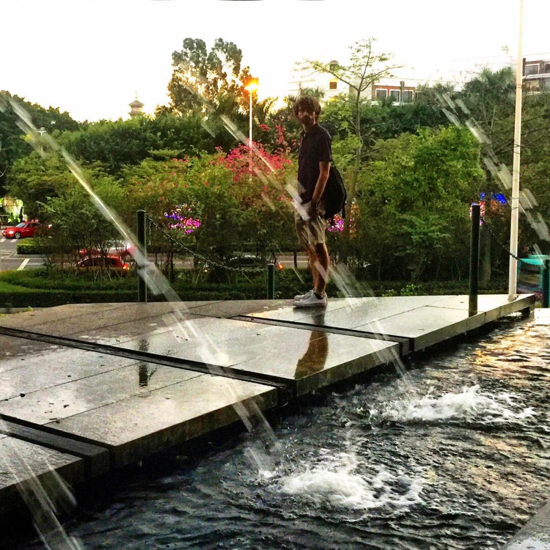 A man stands on some stones over some water. Fountains spray water in front of him. In the background are trees