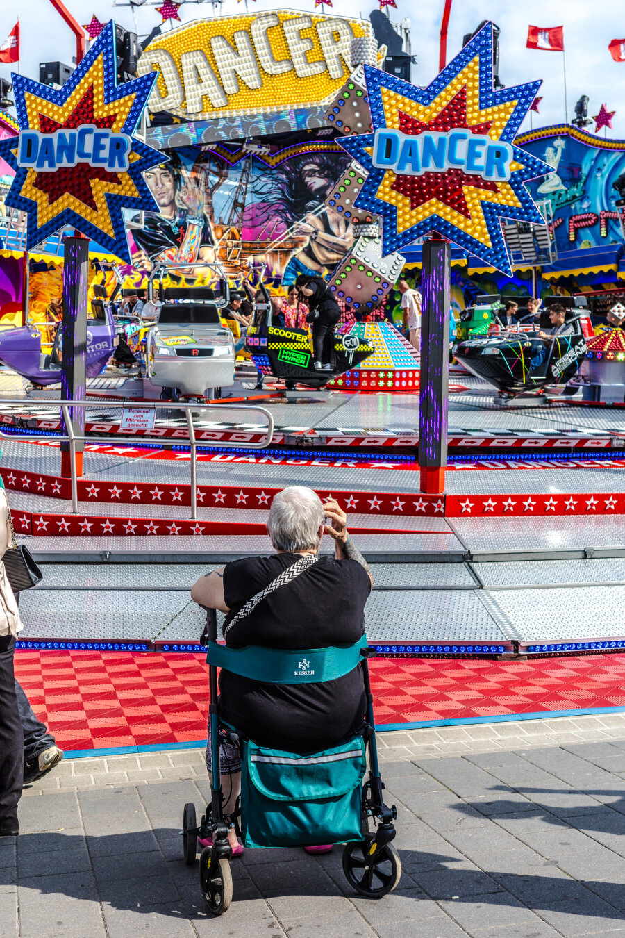 Im Vordergrund sitzt eine Person mit grauen Haaren in einem türkisfarbenen Rollator und blickt auf ein Fahrgeschäft. Das Fahrgeschäft trägt den Namen »Dancer«, der auf zwei großen, sternförmigen Schildern in blauer Schrift auf gelbem Grund steht. Die Kulisse ist bunt gestaltet und zeigt Comicfiguren und Lichtelemente. Mehrere Fahrkabinen mit Menschen bewegen sich auf der Plattform. Der Boden vor dem Fahrgeschäft ist mit einem rot-weißen Muster gestaltet. Die Szene spielt auf dem Hamburger Dom, einem bekannten Volksfest.