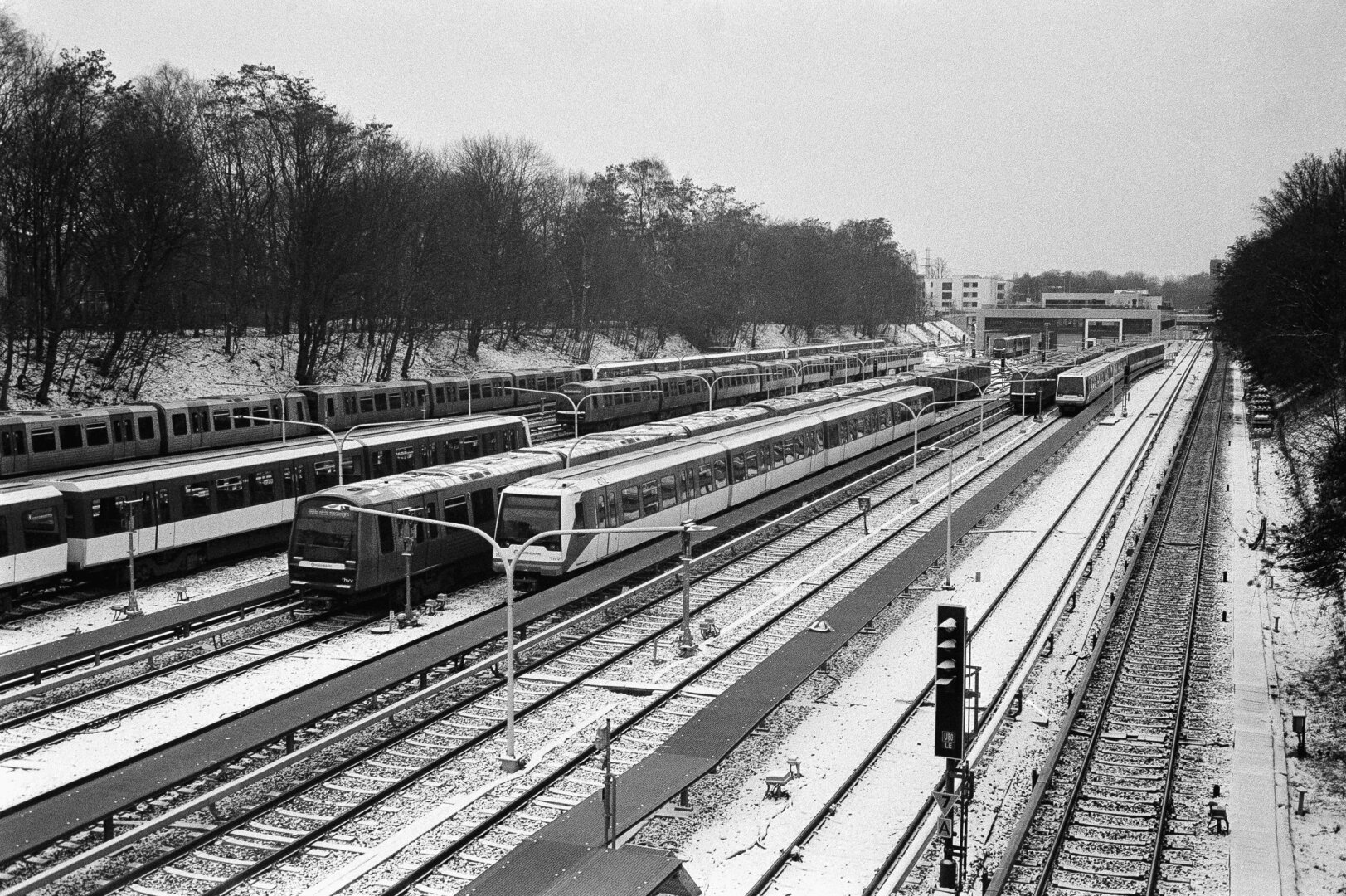 Schwarz-weißes Foto einer U-Bahn-Anlage mit mehreren Gleisen und Zügen. Die Bahnsteige und Gleise sind von einer dünnen Schneeschicht bedeckt. Links und rechts verlaufen parallele Gleise, auf denen U-Bahn-Züge stehen oder fahren. Im Hintergrund sind Bäume ohne Laub und einige Gebäude zu sehen. Die Szene wirkt ruhig und winterlich.