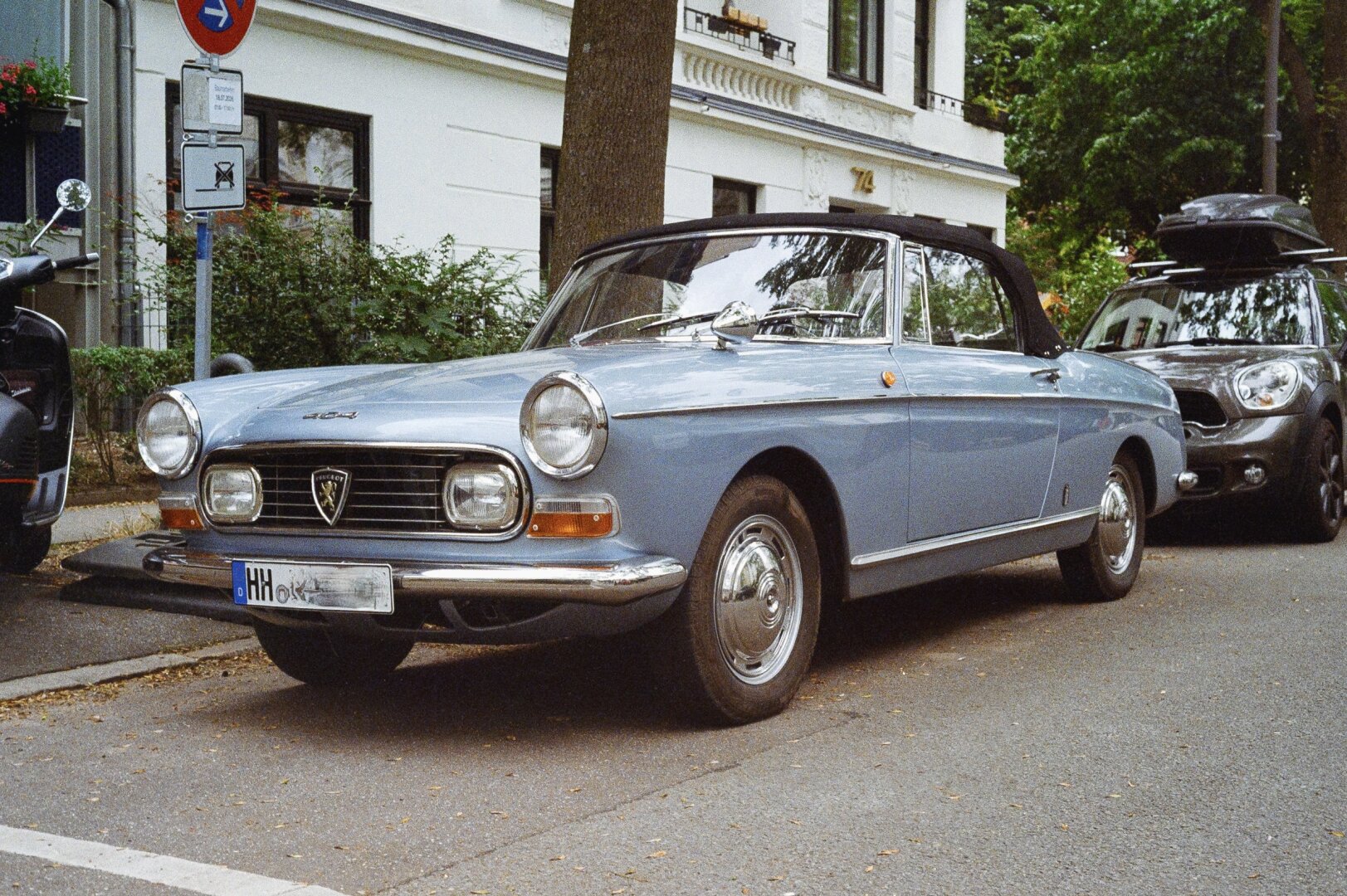 Ein hellblauer Oldtimer mit glänzenden Chromdetails steht am Straßenrand vor einem modernen Fahrzeug. Das Cabriolet besitzt einen schwarzen Stoffdachbezug und runde Scheinwerfer. Die Karosserie zeigt klare Linien und einen markanten Kühlergrill mit einem Emblem. Im Hintergrund sind ein weißes Gebäude mit großen Fenstern, Bäume und Straßenschilder zu sehen.