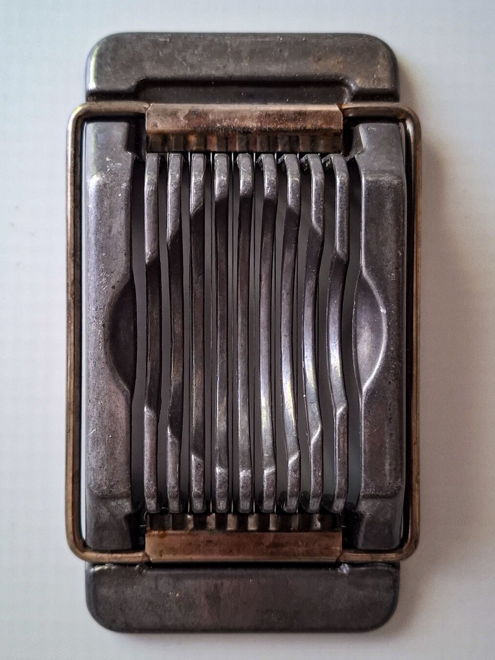 Nahaufnahme eines alten Eierschneiders aus Metall auf hellem Grund. Die klappbare Vorrichtung mit feinen Schneidedrähten ist geschlossen und liegt in den länglichen Rillen der Basisplatte. Das graue Metall ist matt, angelaufen und zeigt deutliche Gebrauchsspuren.