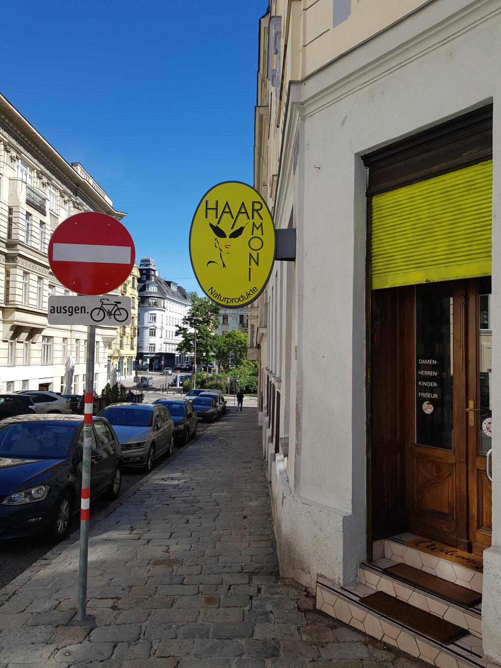 Straßenszene bei Sonne. Rechts ein Friseursalon mit der Tür-Aufschrift „DAMEN- HERREN- KINDER- FRISEUR“. Das gelbe, ovale Ladenschild zeigt ein Frauengesicht und den Text „HAAR MONI Naturprodukte“. Links stehen geparkte Autos und ein rotes „Einfahrt verboten“-Schild (Fahrräder ausgenommen).