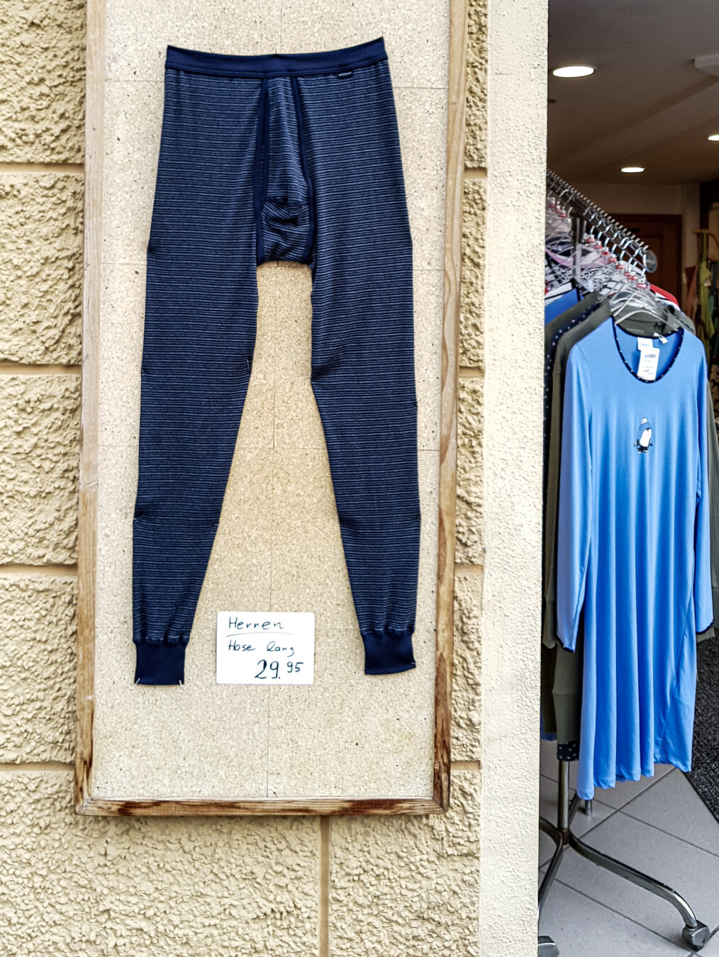An einer beigen Schautafel hängt eine lange, dunkelblau gestreifte Herrenunterhose. Ein handbeschriftetes Schild darunter weist sie als "Herren Hose lang" für 29,95 aus. Rechts daneben steht ein Kleiderständer mit einem blauen Nachthemd im Eingangsbereich des Ladens.