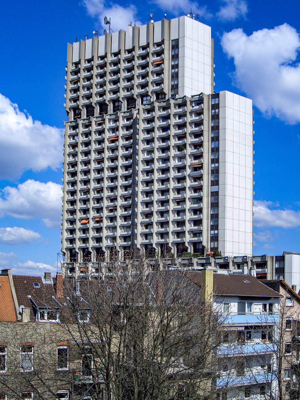 Ein massives Wohnhochhaus im brutalistischen Stil ragt dominant in den blauen, leicht bewölkten Himmel. Die Fassade besteht aus unzähligen, wabenartig angeordneten Betonbalkonen, die dem Gebäude eine markante Struktur verleihen. Im starken Kontrast dazu stehen im Vordergrund niedrige Altbauten mit traditionellen Ziegeldächern und kahle Bäume, die von dem gigantischen Komplex weit überragt werden.