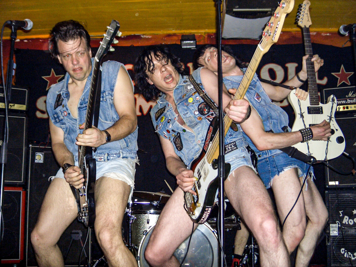 Energischer Auftritt der „Rock'n'Roll Stormtroopers“ am 28.05.2005 im JUZ Mannheim. Drei Musiker performen wild an Gitarren und Bass. Ihr Markenzeichen: ein auffälliger Einheitslook aus mit Patches besetzten Jeanswesten und extrem kurzen Jeans-Hotpants vor einem schwarzen Banner.