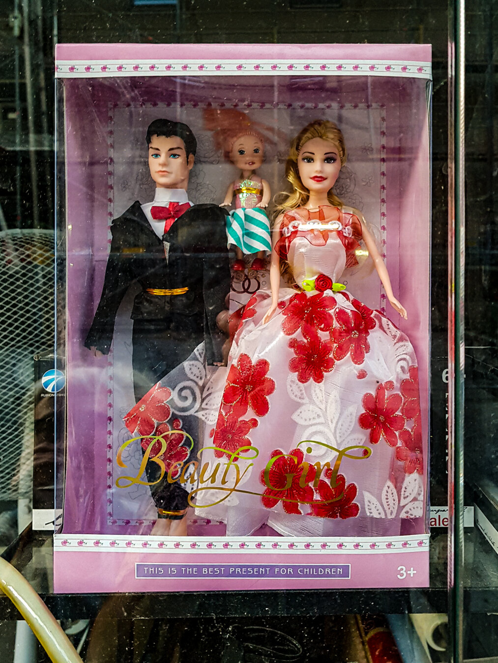Ein Puppen-Set in einer rosafarbenen Schachtel, sichtbar hinter einer Glasscheibe. Es enthält eine blonde Frau in einem weißen Kleid mit roten Blumen, einen Mann im Smoking und eine kleinere rothaarige Puppe, die ein Geschenk hält. Die Aufschrift lautet "Beauty Girl" und "THIS IS THE BEST PRESENT FOR CHILDREN".