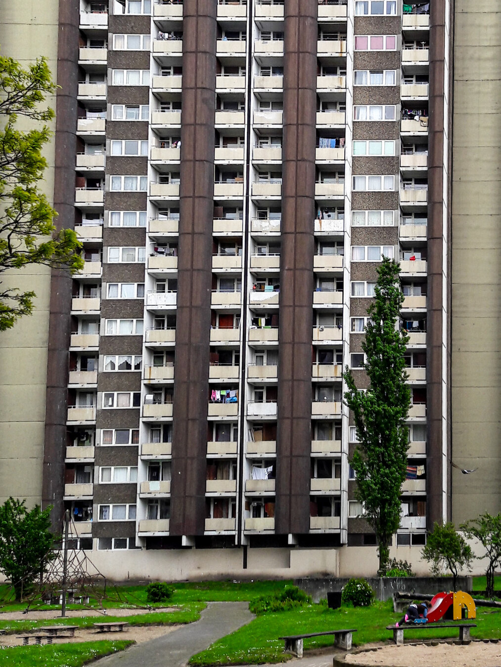 Frontalansicht eines massiven Wohnhochhauses mit grauer Betonfassade, gegliedert durch vertikale dunkle Säulen und zahlreiche Balkone. Auf einigen hängt Wäsche. Im Vordergrund liegt eine Grünfläche mit Bäumen, einem Weg und einem Spielplatz mit roter Rutsche und Klettergerüst.