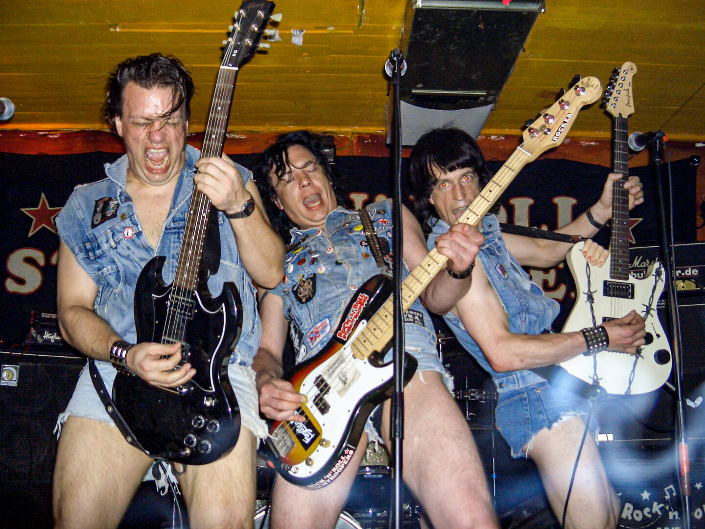 Energischer Auftritt der „Rock'n'Roll Stormtroopers“ am 28.05.2005 im JUZ Mannheim. Drei Musiker performen wild an Gitarren und Bass. Ihr Markenzeichen: ein auffälliger Einheitslook aus mit Patches besetzten Jeanswesten und extrem kurzen Jeans-Hotpants vor einem schwarzen Banner.