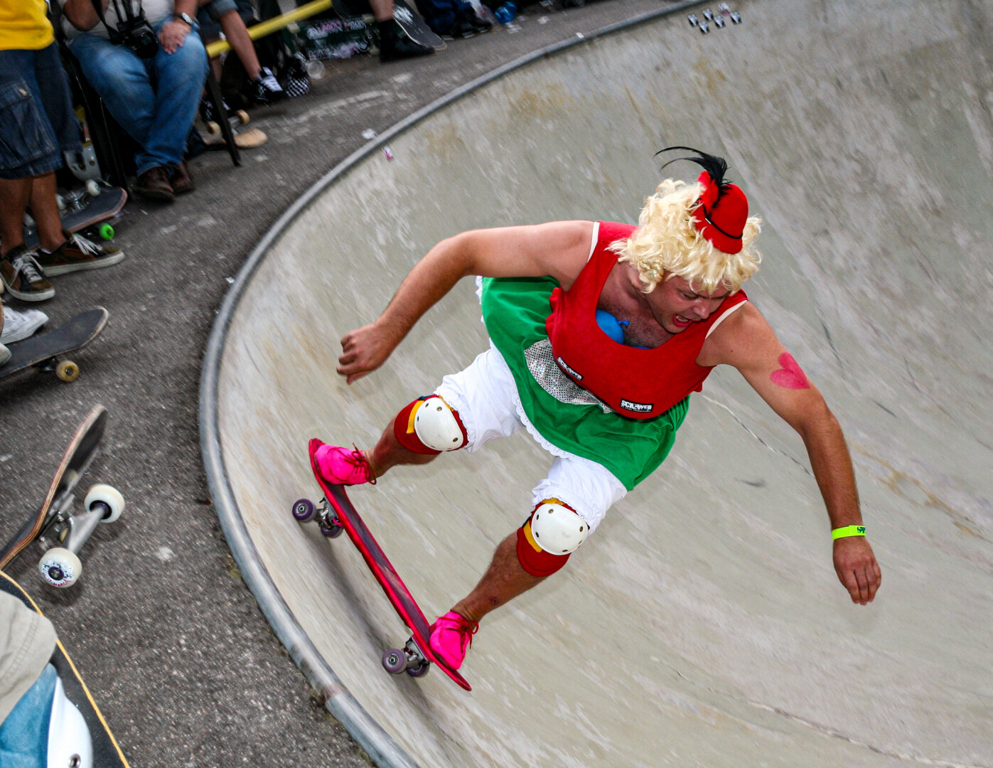 Ein Mann in einem exzentrischen Kostüm fährt dynamisch auf einem roten Skateboard durch eine Beton-Bowl. Er trägt eine blonde Lockenperücke, einen kleinen roten Hut, ein rotes Top, einen grünen Rock über weißen Shorts sowie Knieschoner und pinke Schuhe. Am oberen Rand stehen Zuschauer.