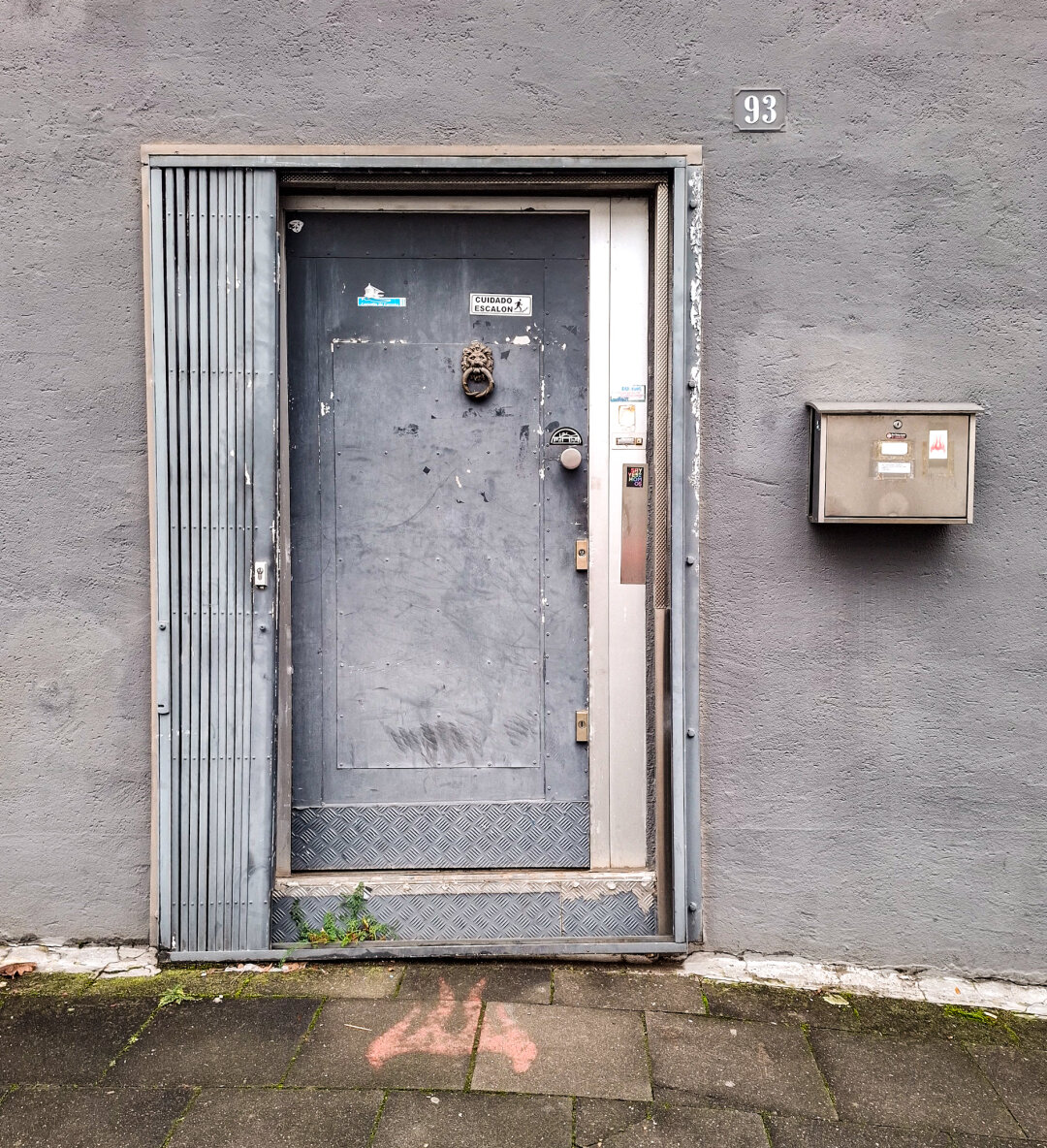 Eine verwitterte, graue Metalltür in einer rauen Fassade, markiert mit der Hausnummer 93. Rechts hängt ein silberner Briefkasten, links ein zurückgeschobenes Gitter. Details wie ein Löwenkopf-Klopfer, Sticker, Riffelblech am Boden und ein rotes Graffiti auf dem Gehweg prägen das Bild.
