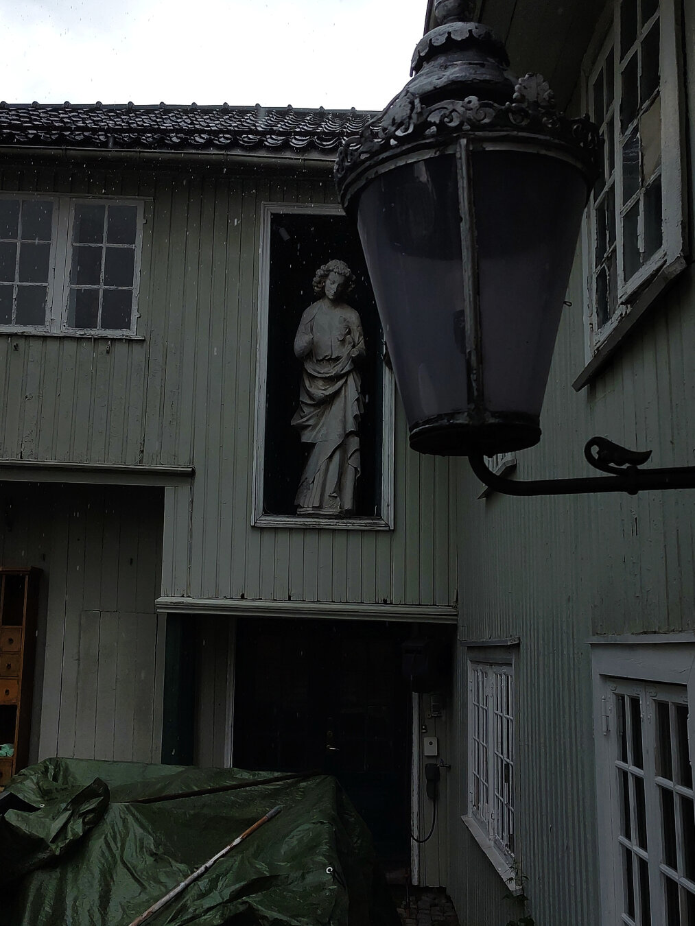 Скульптура Девы Марии (?) в стене старого дома, в котором идет ремонт. У стены, под скульптурой сложены строительные материалы или инструменты. Они накрыты мешковиной. Справа на кадре обшитая деревом стена с окнами. Моросит мелкий дождь