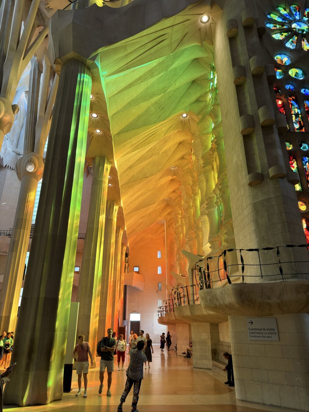 Towering stone columns inside Barcelona’s Sagrada Familia, bathed in green-to-red stained-glass light, with visitors walking below.