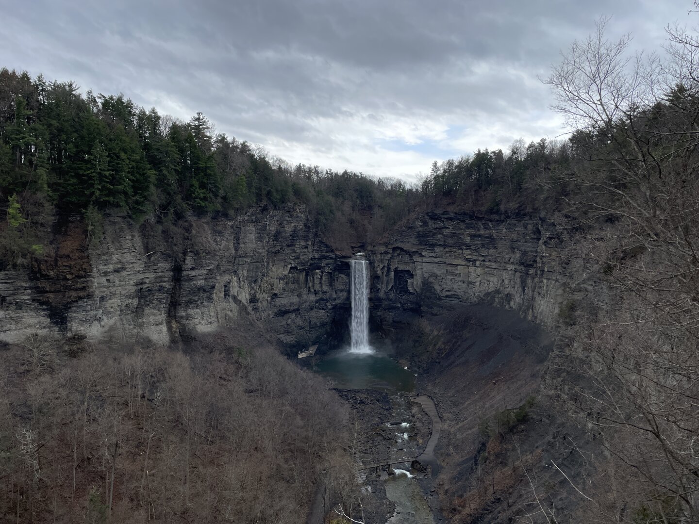 A tall waterfall pours down the wall of a gorge. It is late winter, so many of the trees around the gorge are still bare. The sky is very cloudy.